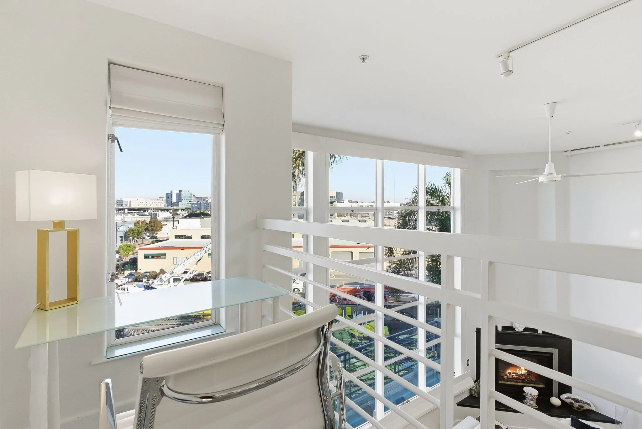 Bright, modern interior of a living space with large windows overlooking an urban landscape, a white desk with a gold and white lamp, a white chair, and a fireplace.