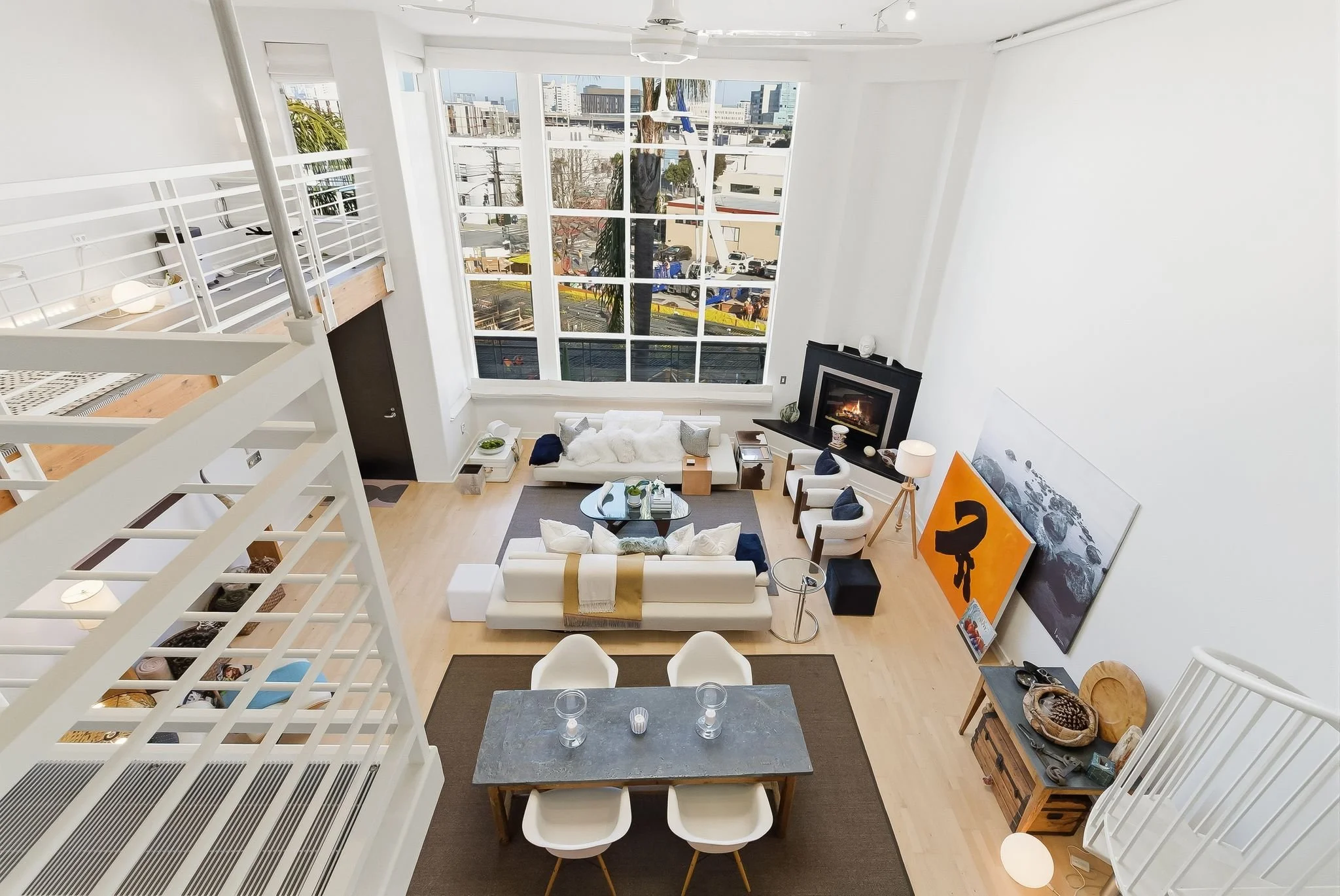 A spacious, modern living room with a high ceiling and large windows, featuring white walls, light wood flooring, a fireplace, a white sofa, two armchairs, a coffee table, a TV, and various decorative items and artwork.