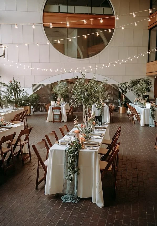 A decorated indoor event space with long tables covered in white tablecloths, adorned with floral centerpieces, greenery, and candles. String lights hang from the ceiling, and there are potted plants and additional tables in the background. The space has modern architecture with large round windows and wooden accents.