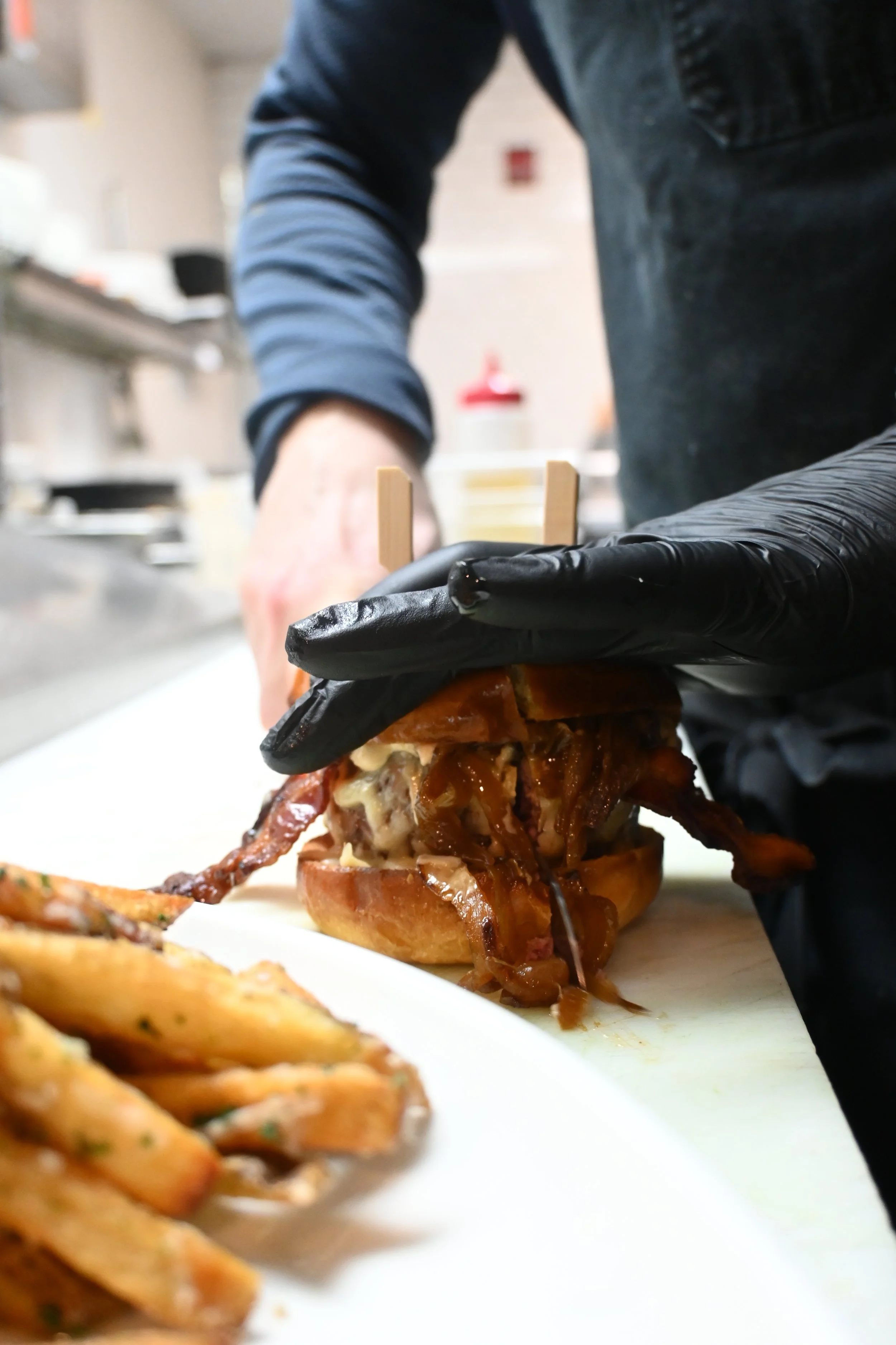 A person wearing black gloves assembling a large, loaded burger with pulled pork and caramelized onions, with a side of fries in the foreground.