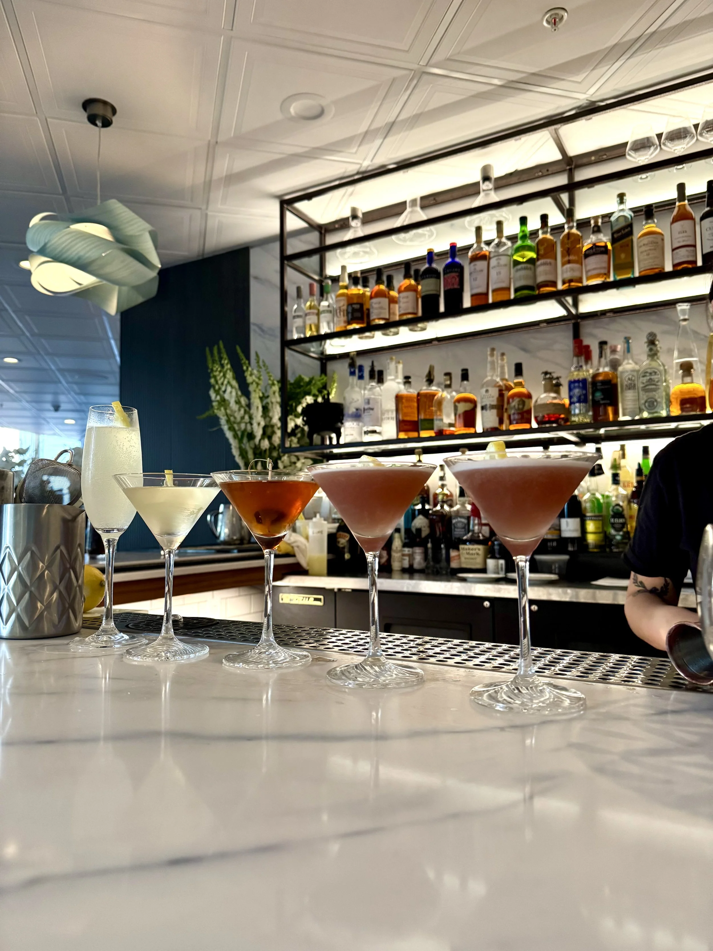 Five cocktails with different colored drinks lined up on a bar counter in front of a backlit liquor shelf.