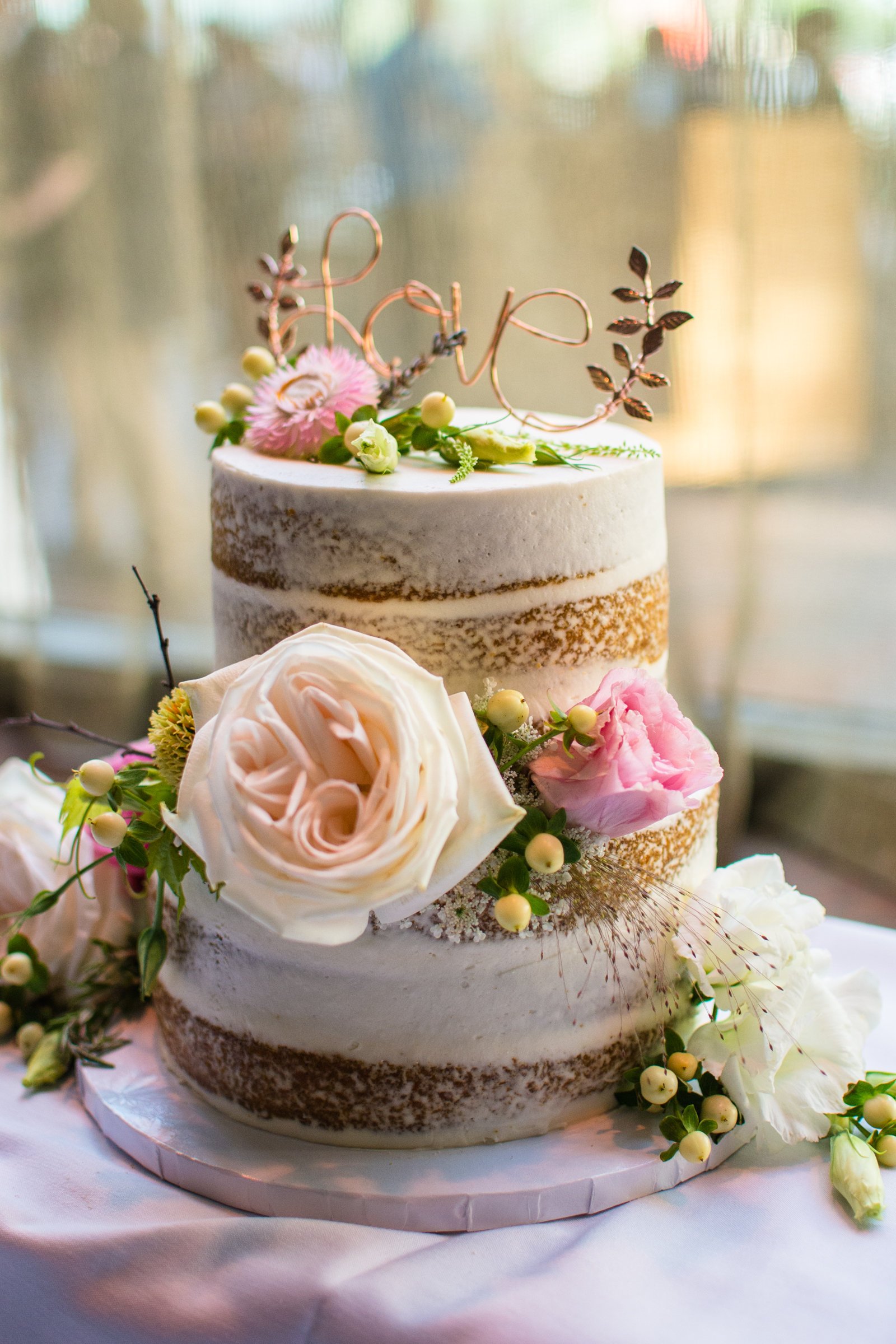 A two-tier wedding cake decorated with fresh flowers and a copper wire topper that spells "love". The cake has a semi-naked frosting style, with the layers partially visible through the thin layer of frosting. The cake is surrounded by additional flowers and greenery on a white tablecloth.