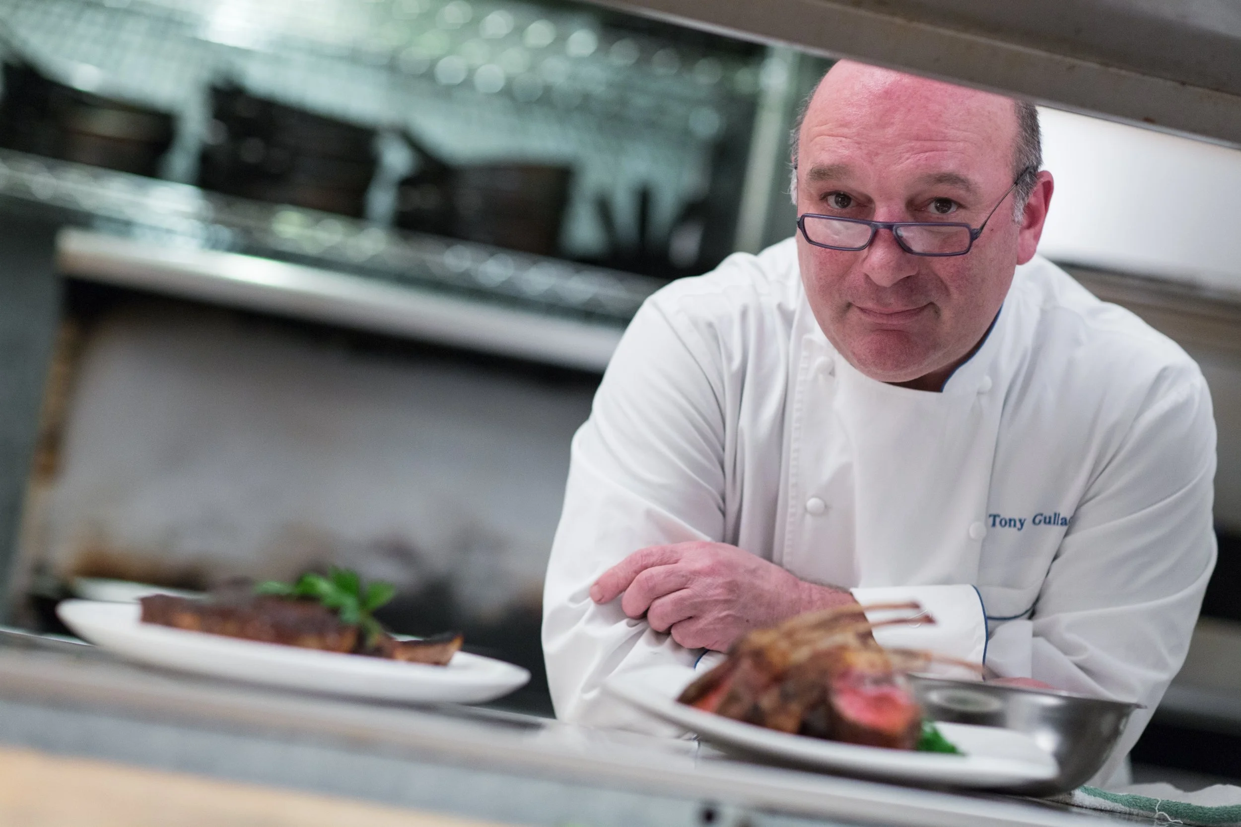 A chef wearing glasses and a white chef's uniform with the name Tony Gullan is leaning over a kitchen counter, with plates of cooked meat dishes in front of him.