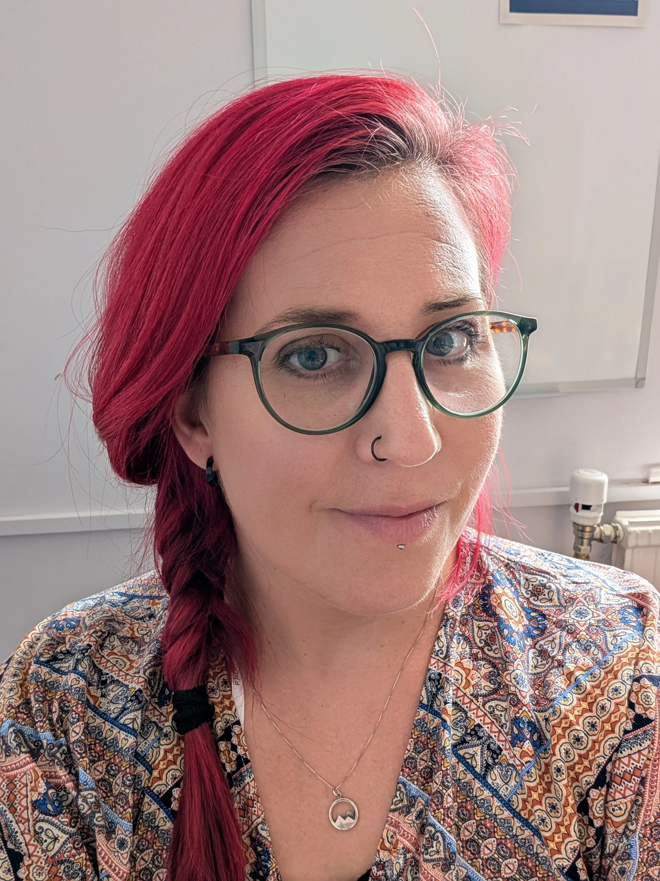 A woman with bright pink hair styled in a braid, wearing glasses, a nose ring, and a necklace, smiling softly indoors.