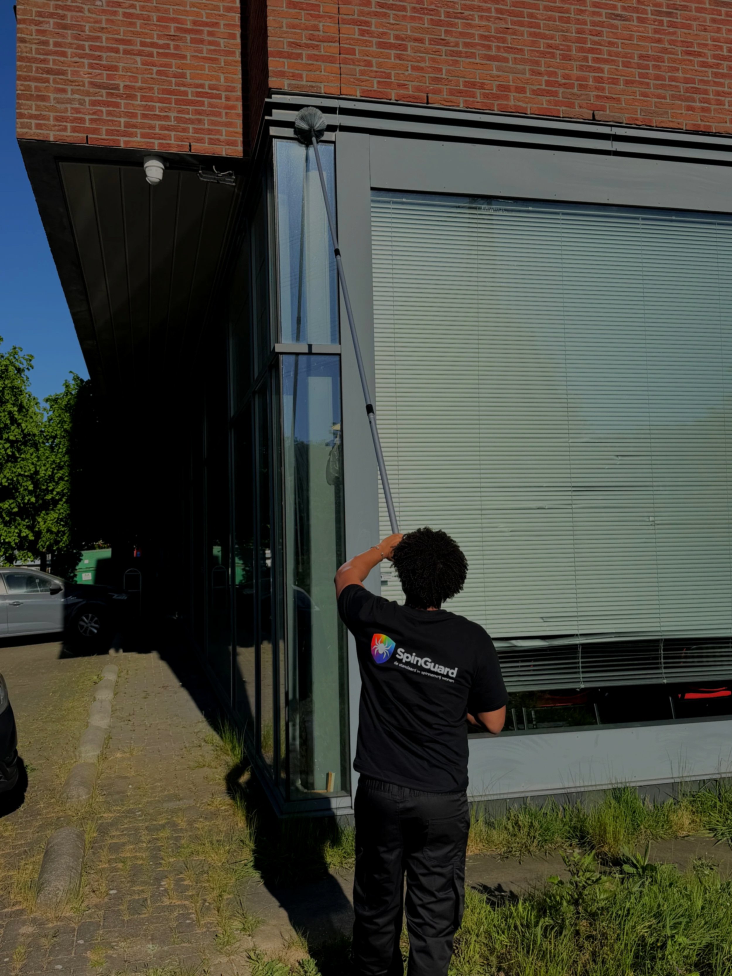 Man met een SpinGuard uniform reinigt een raam met een lange dweil voor een modern gebouw met grote ramen en een rode bakstenen muur onder een heldere blauwe hemel.