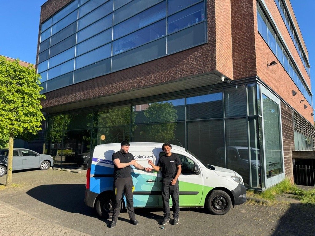 Twee mannen staan buiten een bedrijfspand, de ene overhandigt de andere een sleutel, terwijl ze naast een bedrijfsbus staan.
