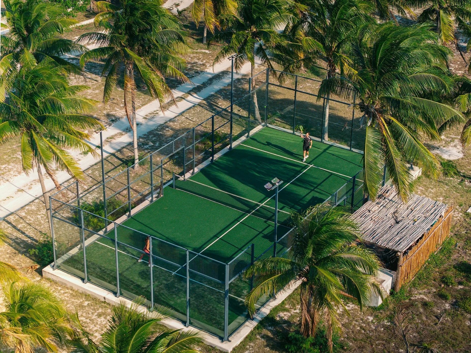 Una cancha de pádel al aire libre rodeada de palmeras, con una persona jugando y otra esperando su turno, en una zona con senderos de tierra.