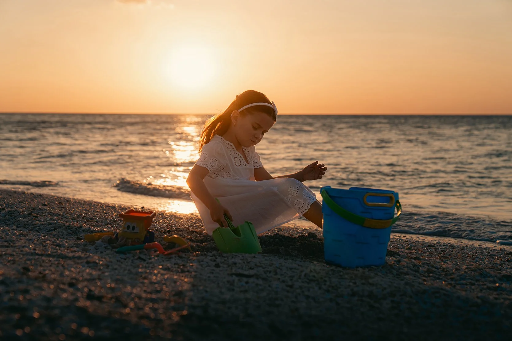 Una niña jugando en la playa al atardecer, con cubetas y juguetes, cerca del mar