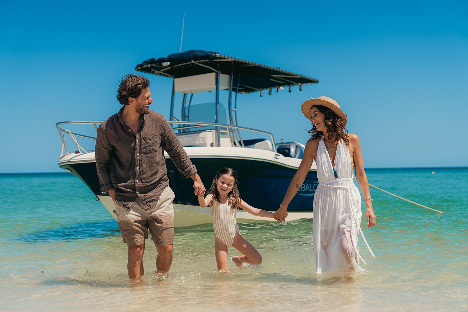 Familia en la playa cerca de un yate, sosteniéndose de las manos y sonriendo bajo el sol.