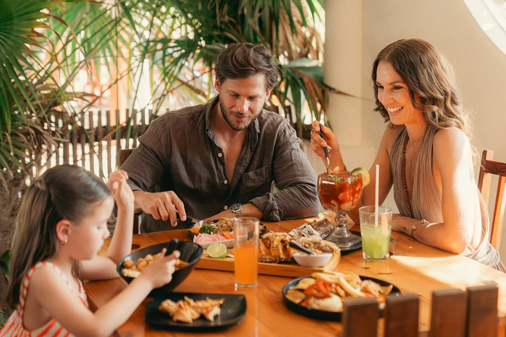 Una familia cenando en un restaurante, con comida y bebidas en la mesa, rodeados de plantas y luz natural en Dunas Yucatán Chuburná México