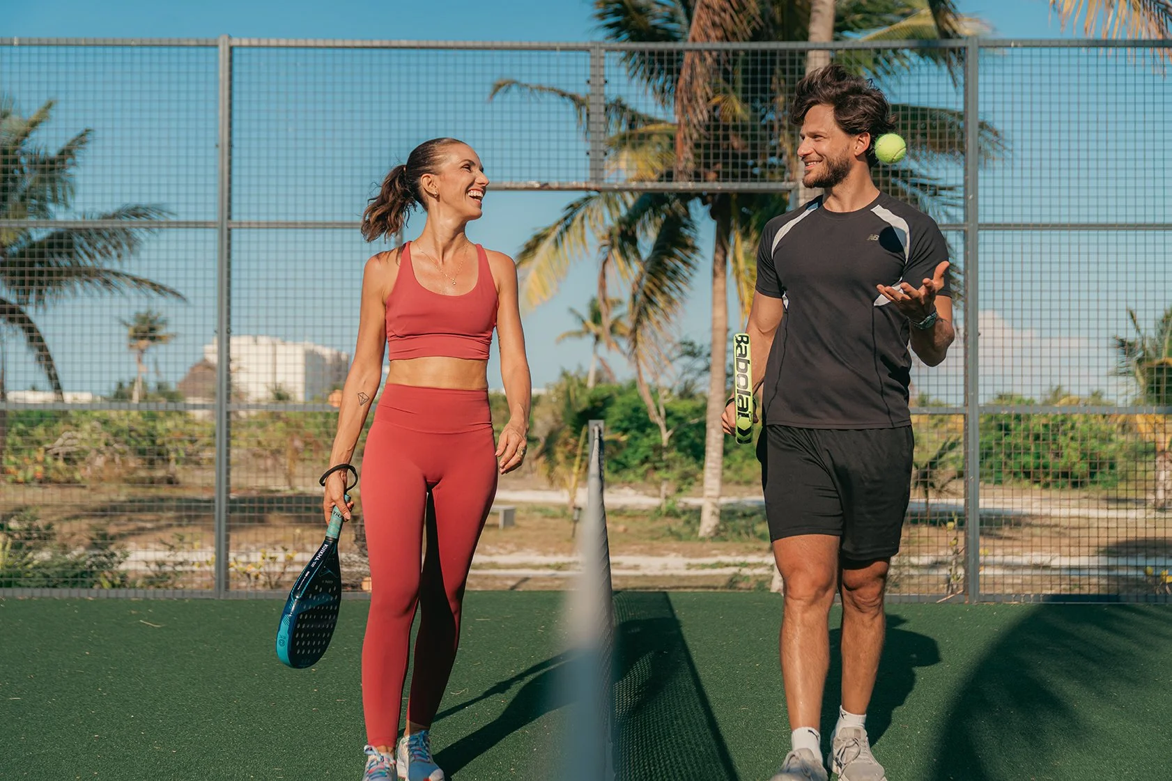 Dos personas jugando tenis en una cancha al aire libre, una mujer con ropa deportiva roja y una mujer con ropa deportiva negra, ambos conversando y sonriendo. La mujer a la izquierda sostiene una raqueta y la otra una pelota de tenis.