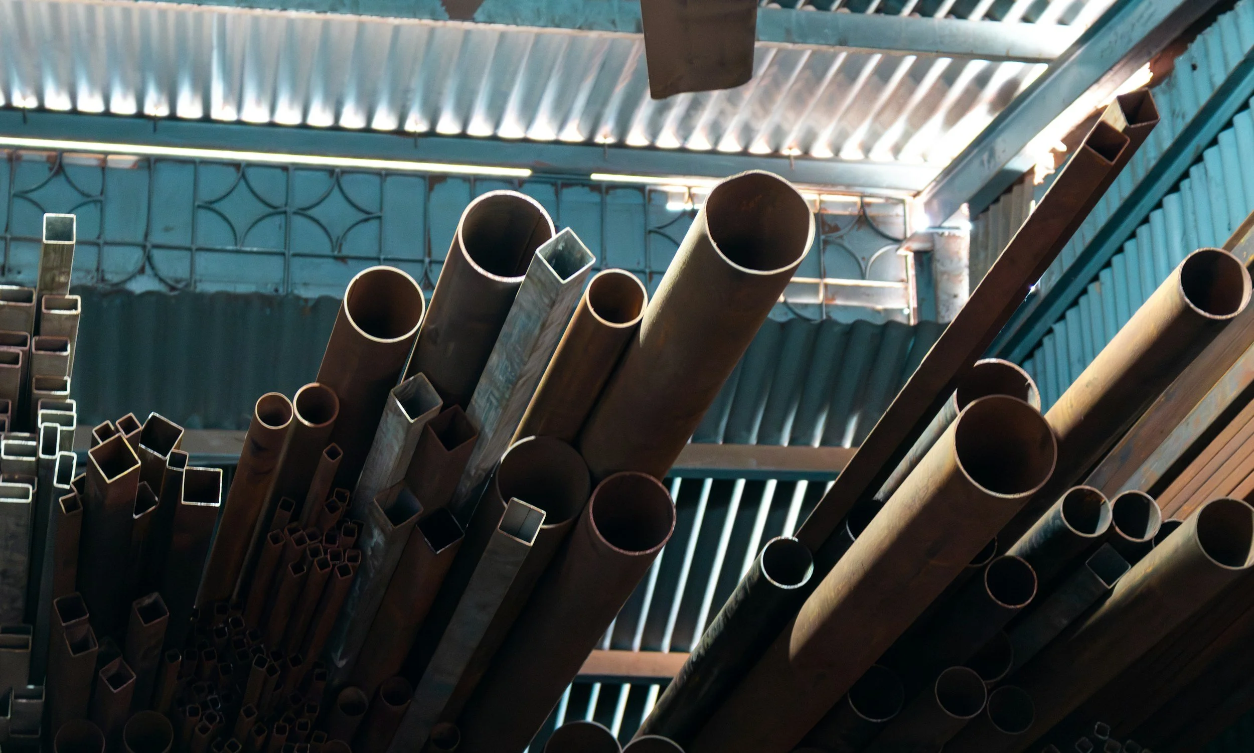 Stacked metal pipes in a warehouse or storage area with corrugated metal roof and walls.