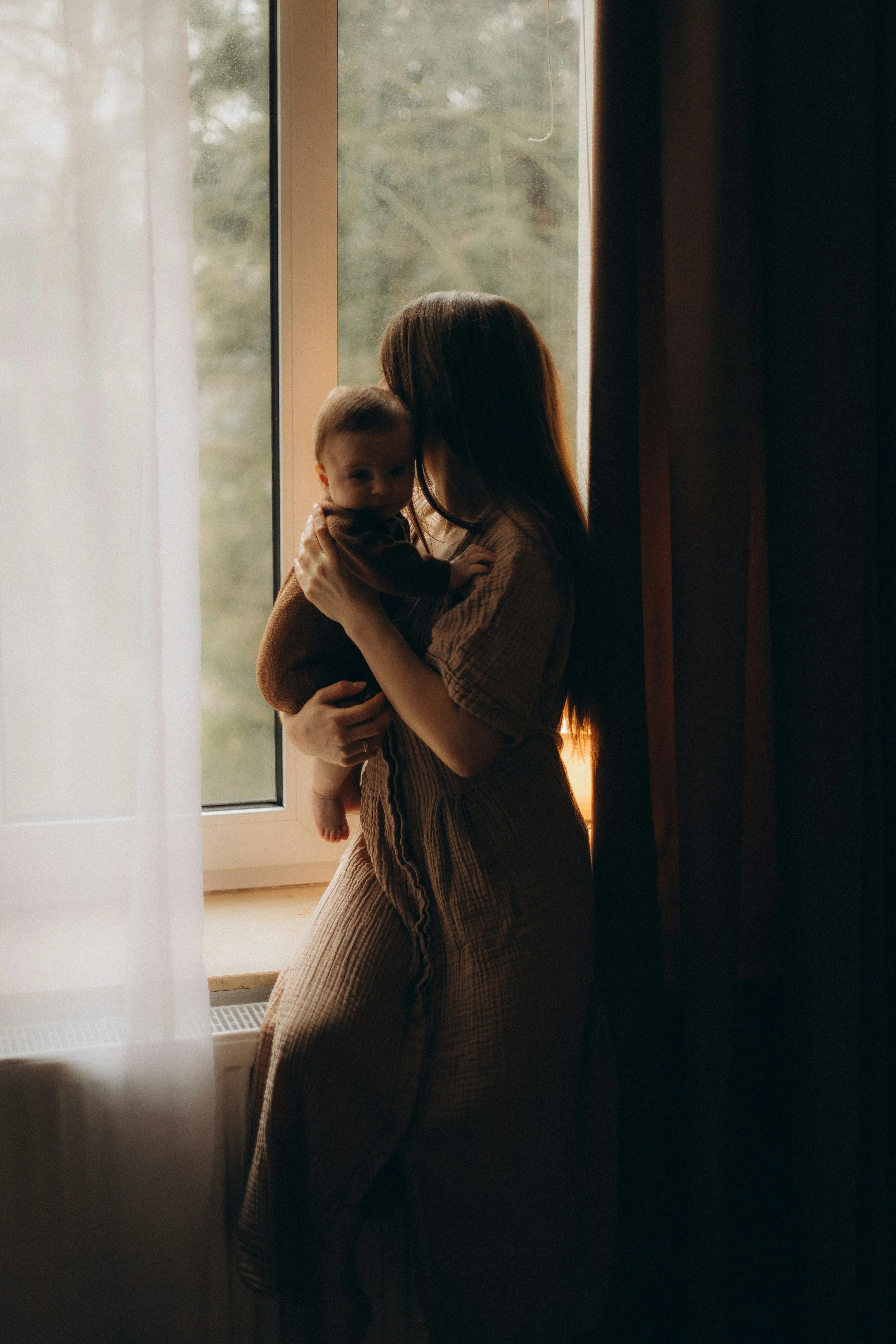 A woman holding a baby near a window with sheer white curtains and dark curtains, soft natural light coming through.
