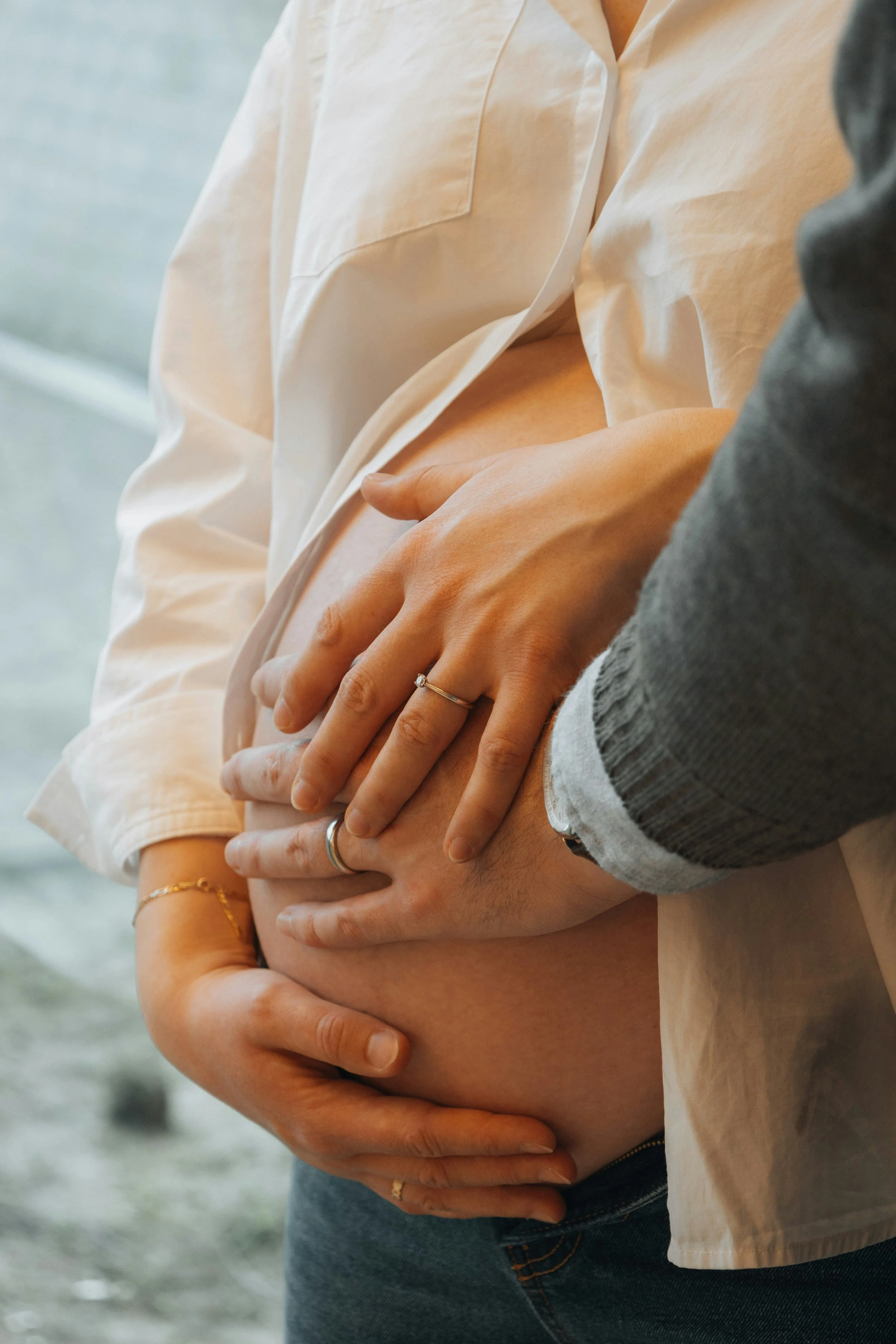 A person in a white shirt and denim jeans is holding their pregnant belly with multiple hands around it, some wearing rings and jewelry.
