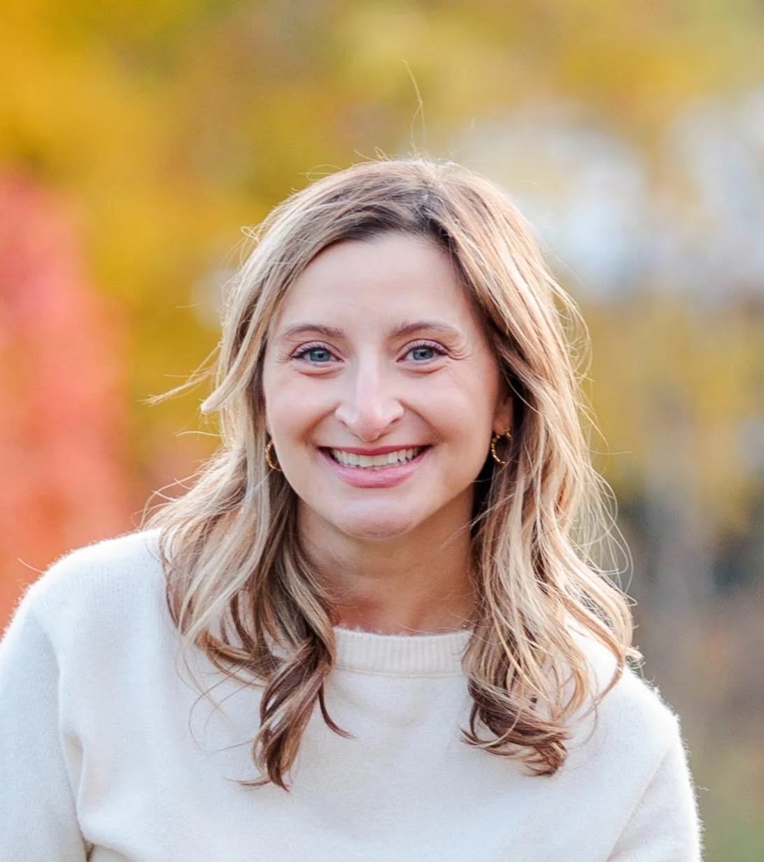 A smiling woman with blonde hair and blue eyes outdoors in autumn with blurred yellow and orange foliage in the background.