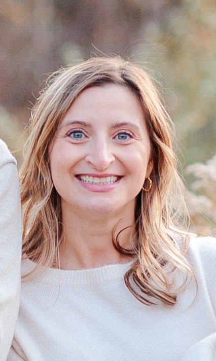 A smiling woman with blue eyes and long blonde hair, wearing a white sweater, outdoors with blurred greenery in the background.