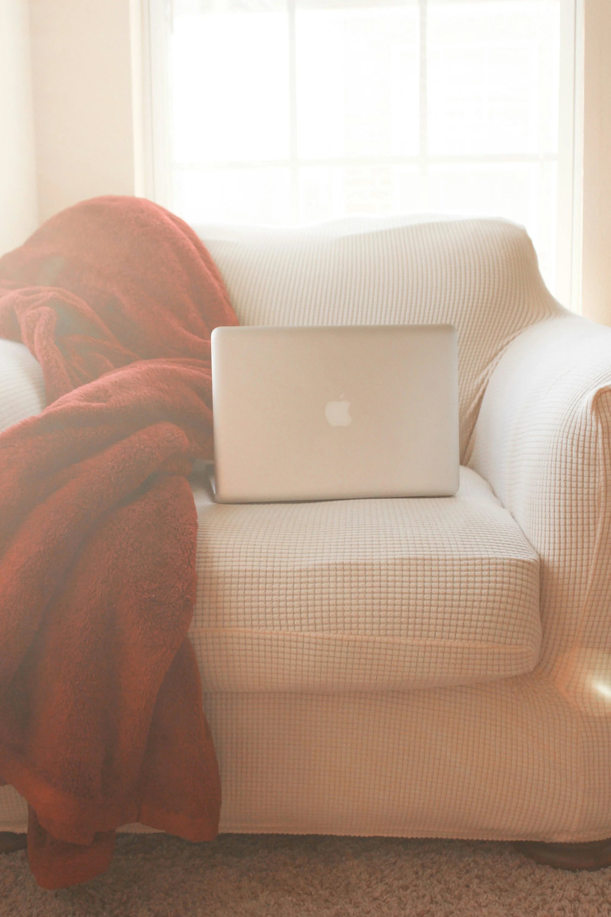 A cozy living room scene with a beige couch, a red blanket, and an open laptop. Light streams in through a window in the background.