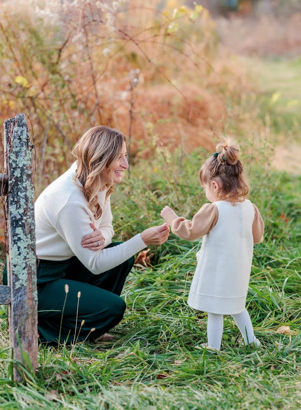 A woman crouching and smiling at a young girl outdoors in a grassy area with autumn foliage, as the girl holds a small object.