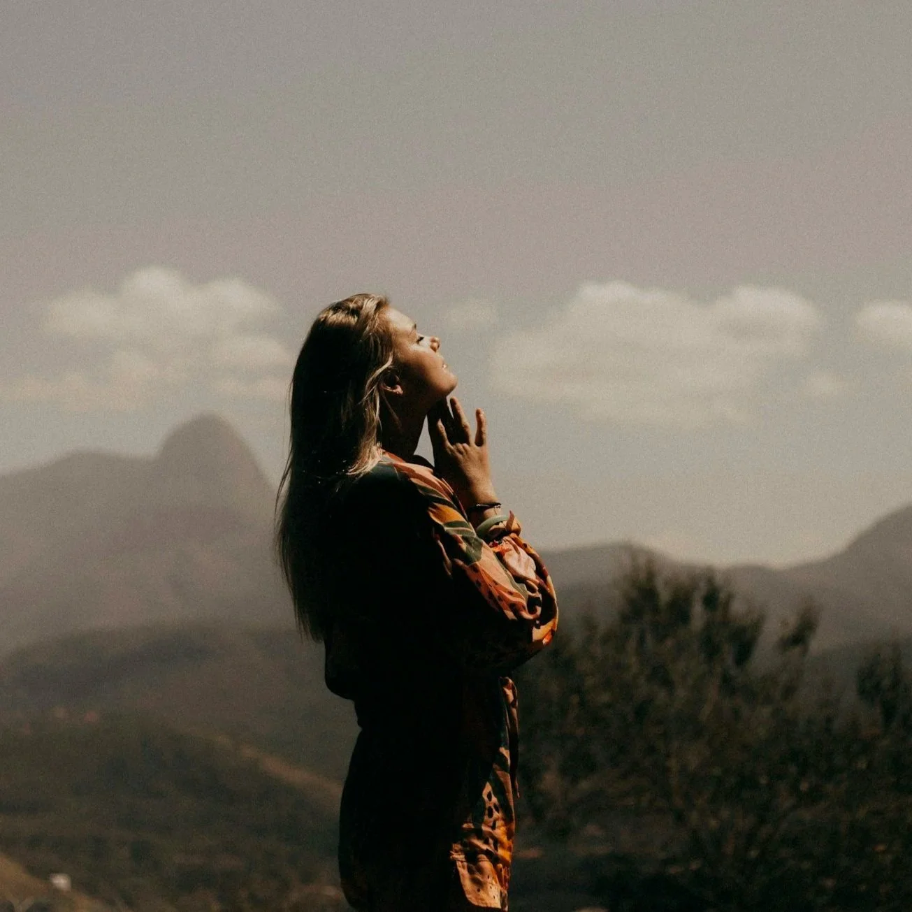 A woman standing outdoors with her eyes closed, facing upward, in a mountainous landscape during daytime.