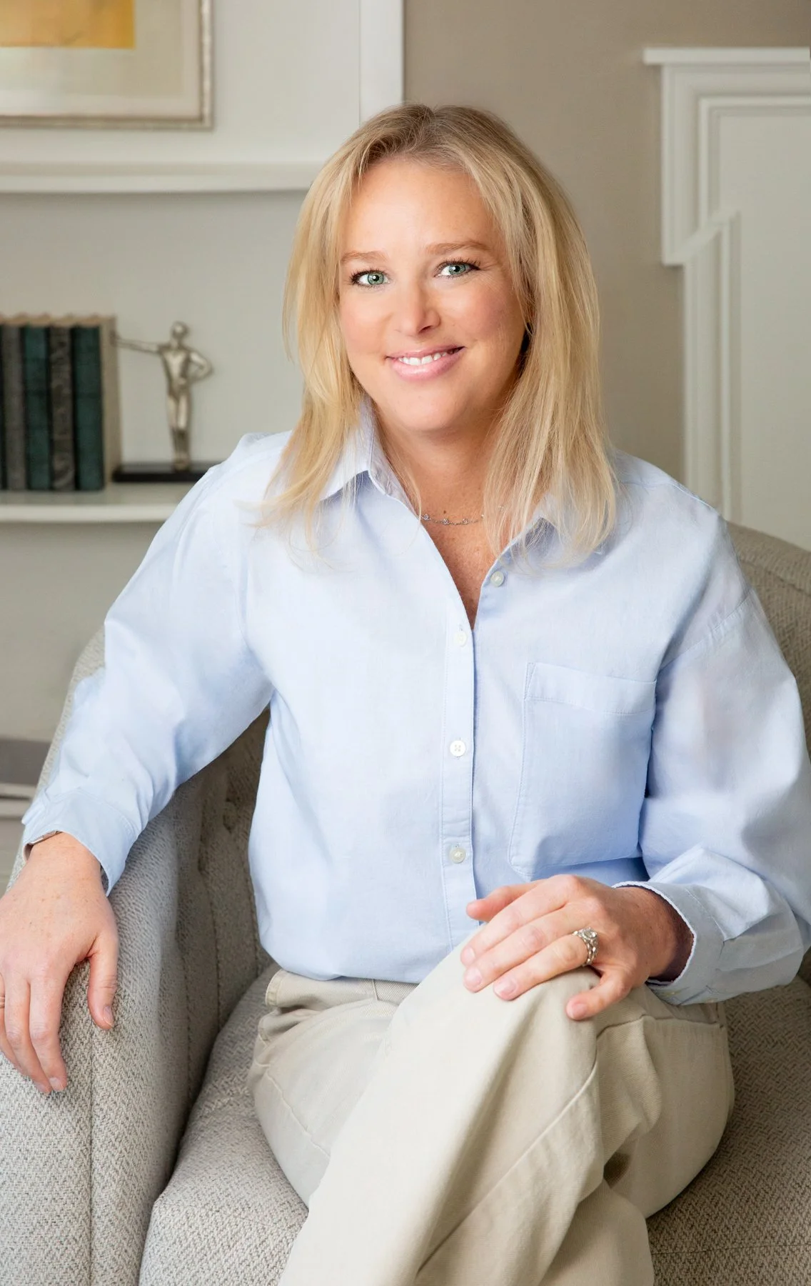 A woman with blonde hair, blue eyes, and a white shirt sitting on a beige couch in a room with light-colored walls, a framed picture, a bookshelf, and a decorative sculpture in the background.