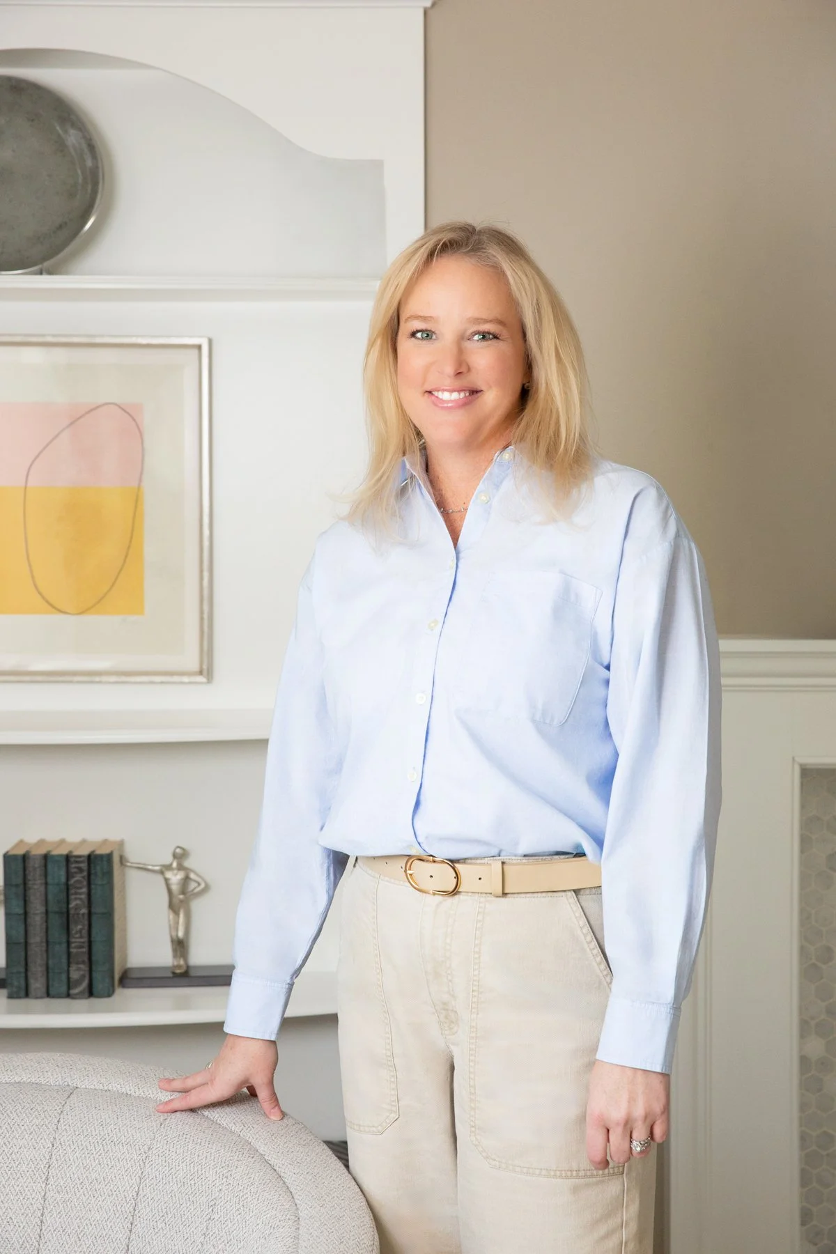 Woman with blonde hair smiling, wearing a light blue button-up shirt and beige pants, standing in a well-decorated room with artwork and books in the background.