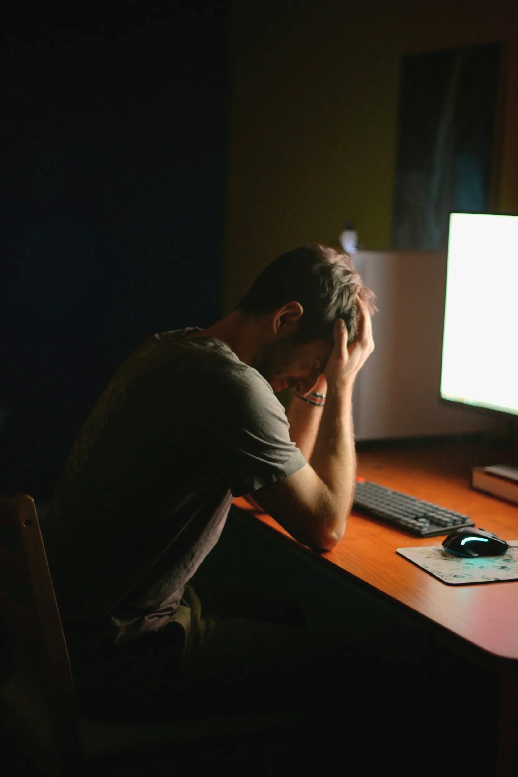 A young man with gray hair sitting at a desk in front of a brightly lit computer monitor, appearing stressed or frustrated with his head in his hands in a dimly lit room.