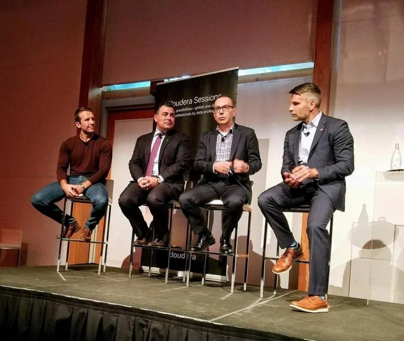 Four men on a stage engaged in a panel discussion, seated on high stools with a black and white banner behind them that reads 'Cloud Expo Session'.