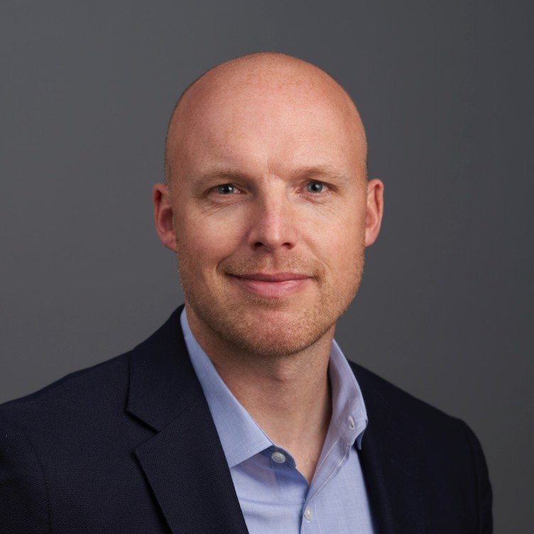 A professional headshot of a bald man with blue eyes, wearing a dark blazer and a light blue collared shirt, against a gray background.