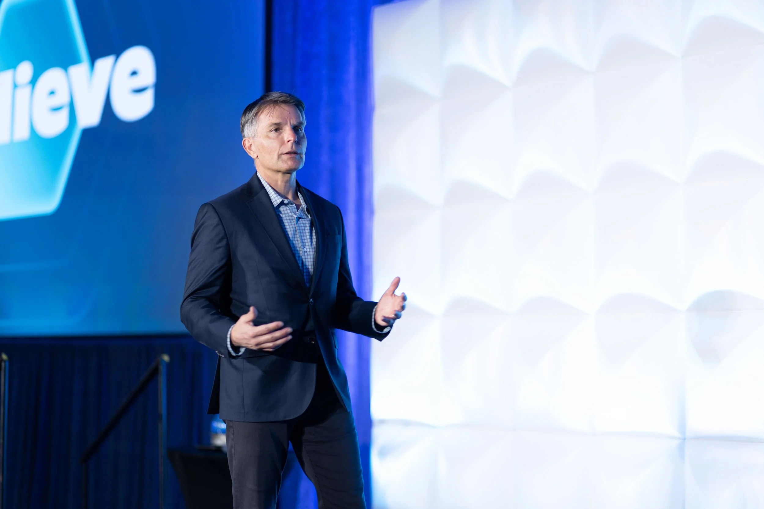 Jeremy Ramsey in a dark suit and checkered shirt giving a presentation on stage, with a blue and white backdrop that has the word 'believe' on it.
