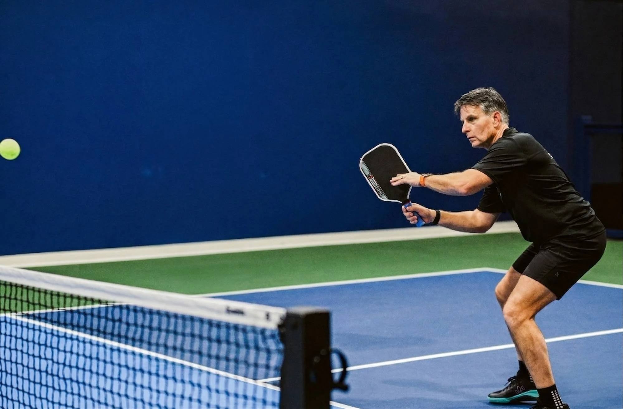 Jeremy Ramsey playing pickleball on an indoor court, wearing a black shirt and black shorts, holding a paddle and preparing to hit a yellow pickleball.