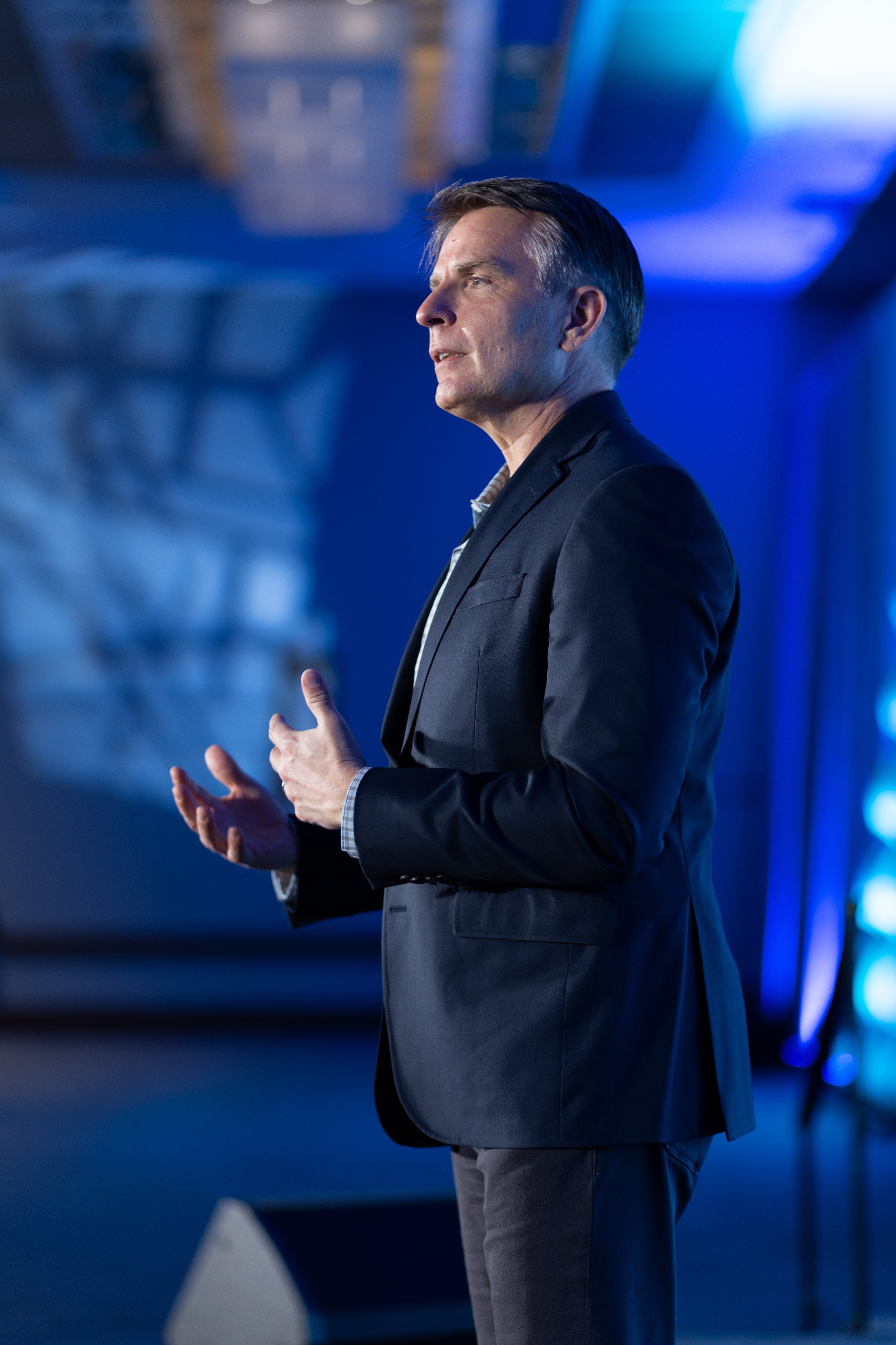 Jeremy Ramsey wearing a dark suit and light shirt is speaking in a dimly lit room with blue lighting, standing with his hands gesturing.