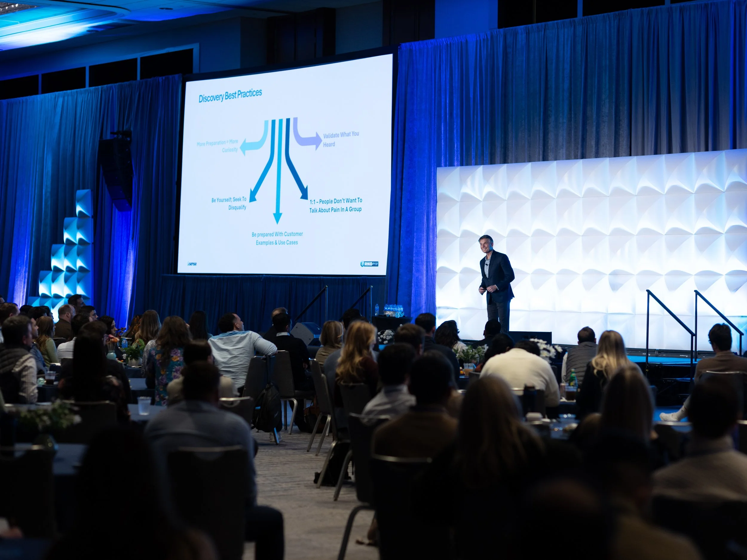 A conference room filled with attendees watching a large presentation on a screen, with a man standing at the front near the stage.