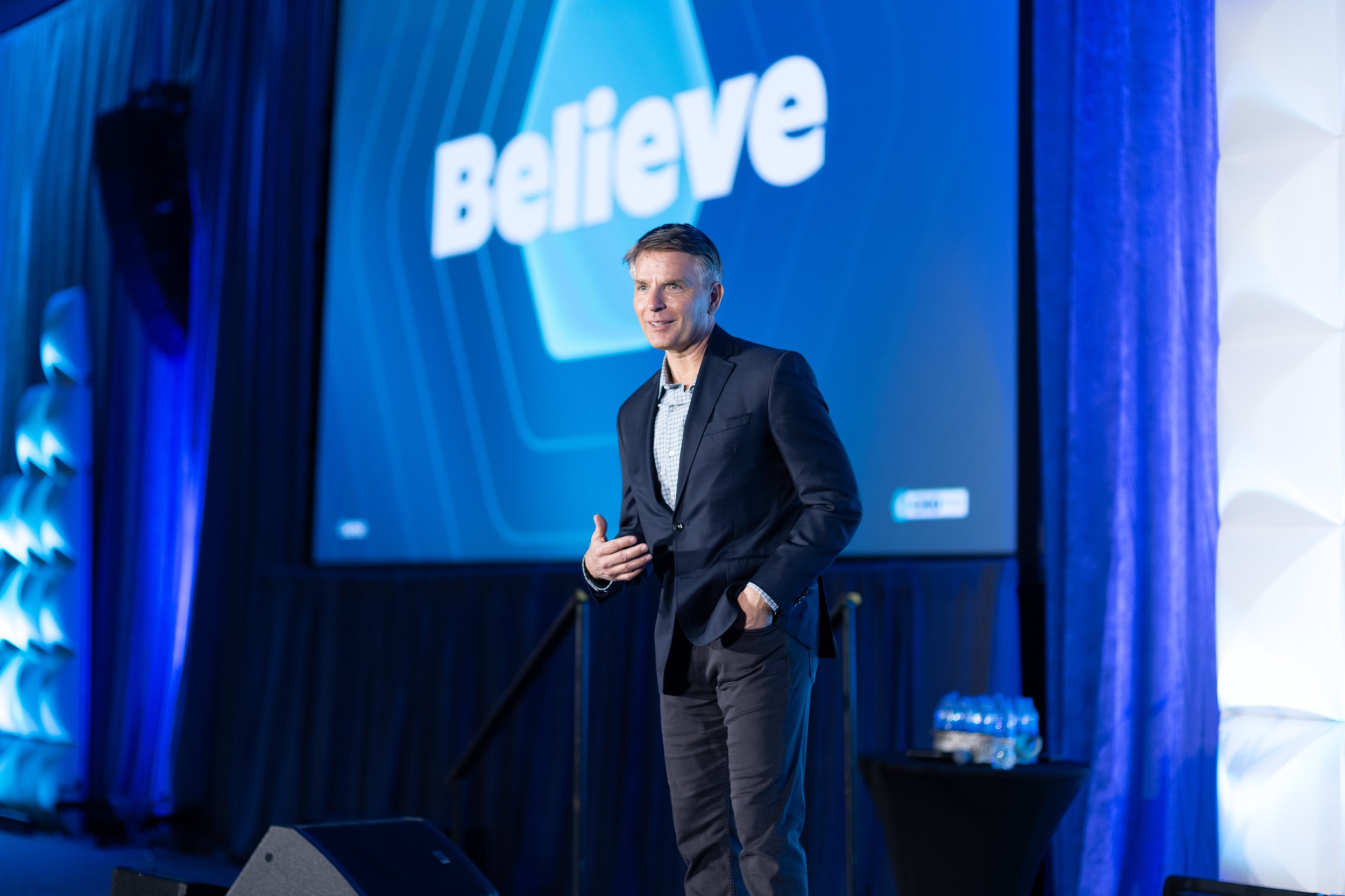 A man in a black suit speaking on stage at a conference, with a large blue screen behind him displaying the word 'Believe' in white text.