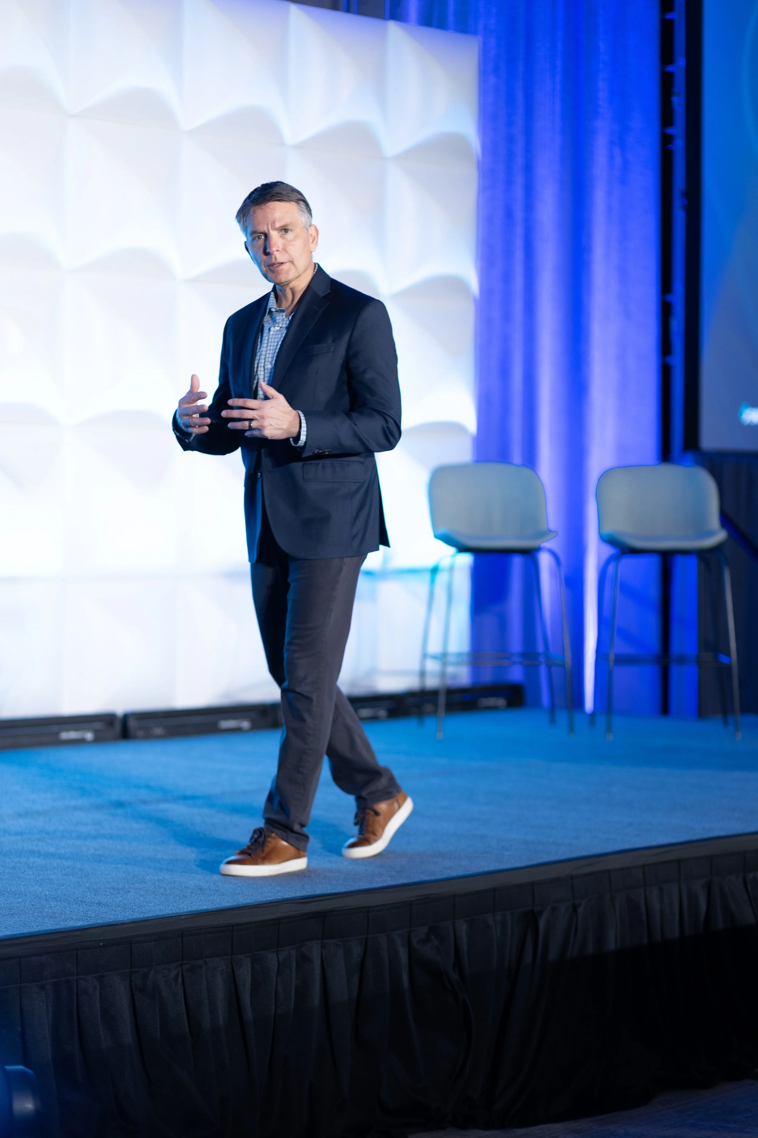 Jeremy Ramsey in a dark suit jacket, checkered shirt, and brown sneakers is standing on a stage, speaking or presenting. The stage has a blue carpet and a backdrop of textured white panels with blue lighting. There are three empty chairs behind him.