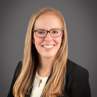 A woman with long blonde hair, wearing glasses, a black blazer, and a white top, smiling against a gray background.