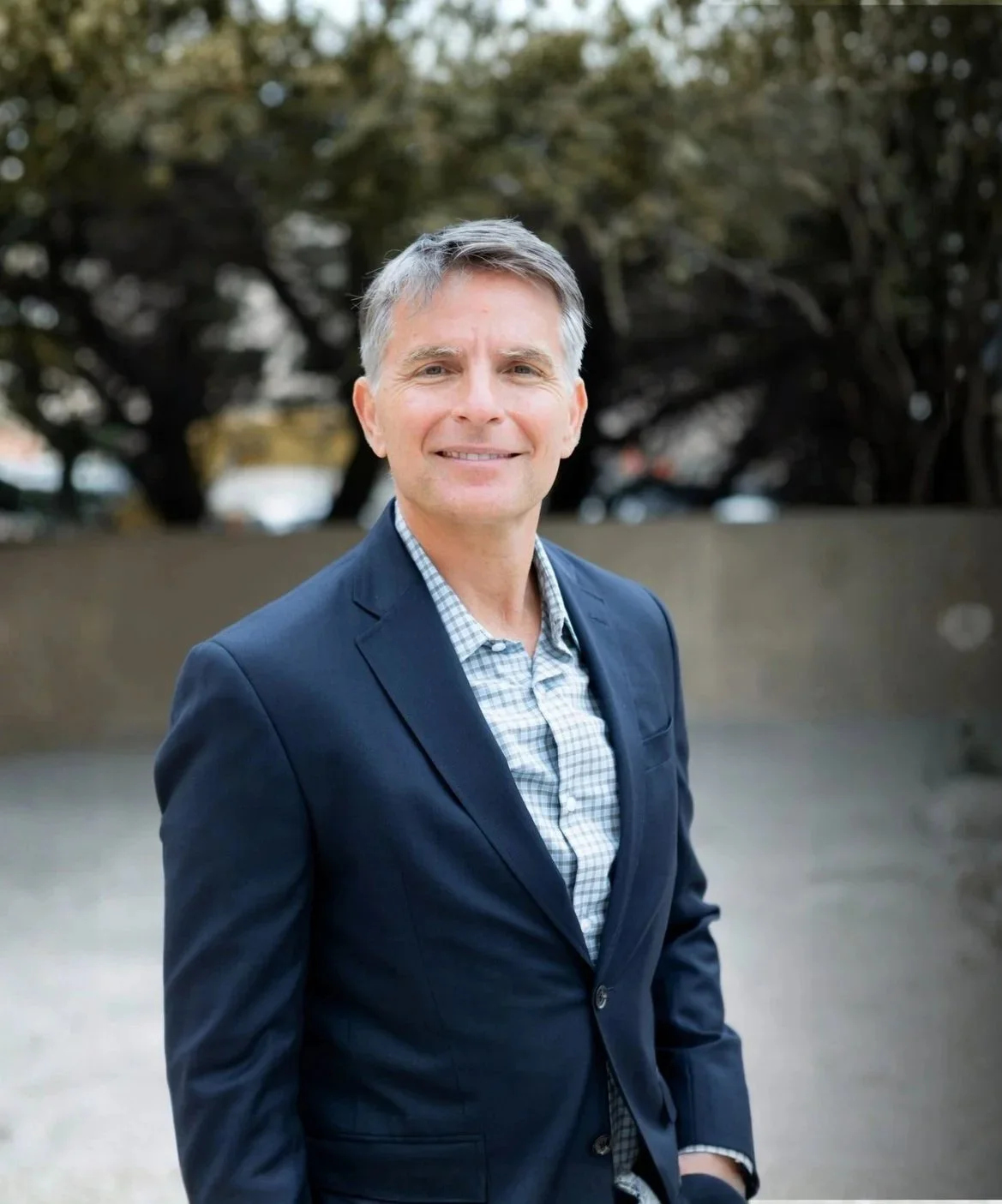 Headshot picture of a middle-aged man in a navy blue blazer and checkered shirt, outdoors with trees and a concrete wall in the background, smiling at the camera.