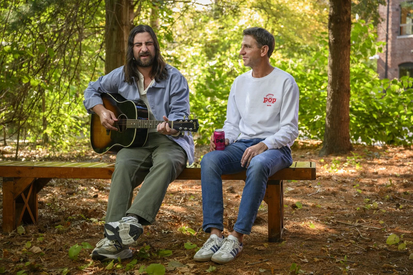 Two men sit on a wooden bench outdoors in a wooded area, one playing an acoustic guitar and singing, the other holding a can of soda and listening.