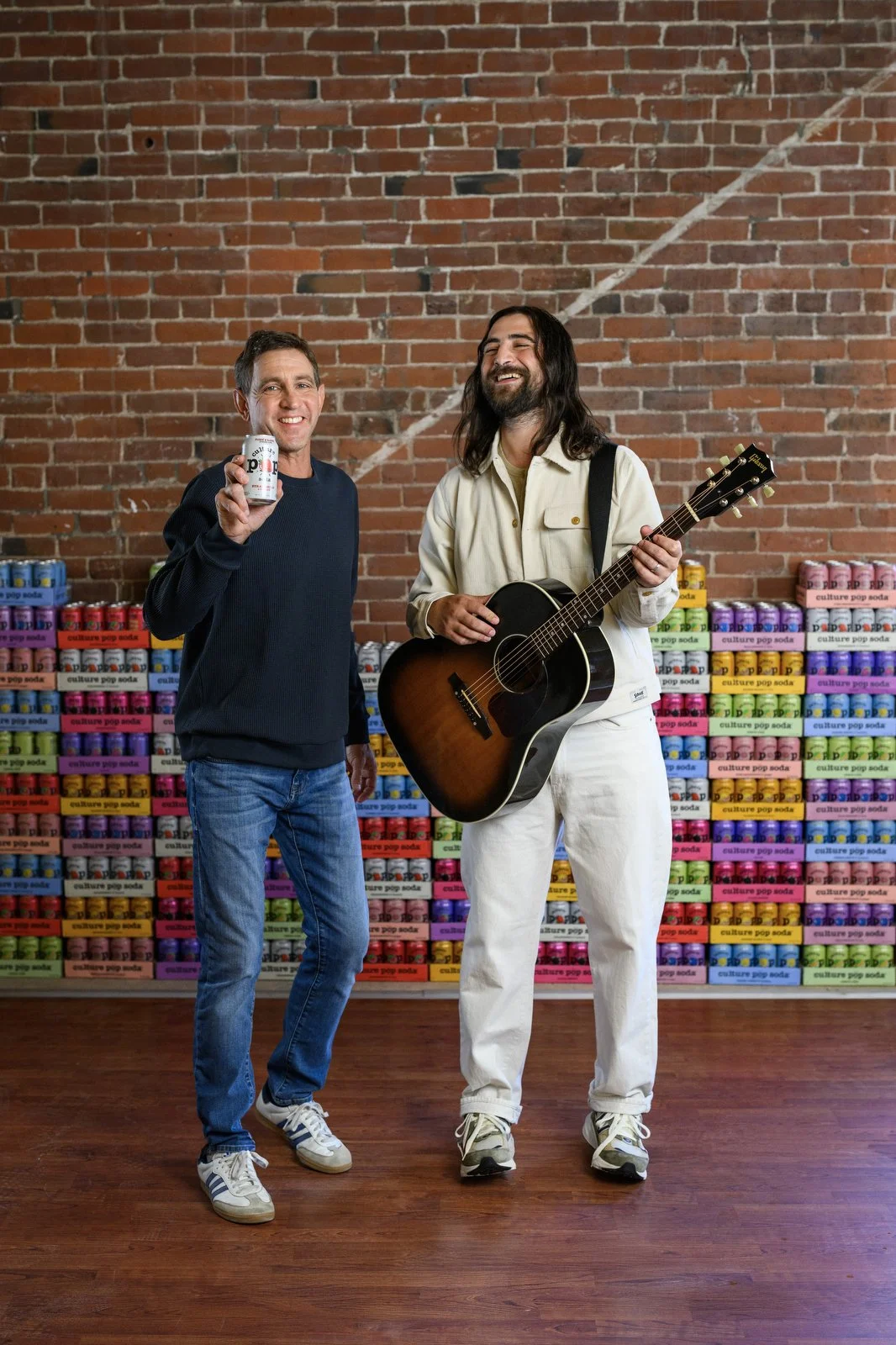 Two men standing in front of a colorful display of cans and boxes of soda against a brick wall. One man is holding a can of pop and smiling at the camera; the other is holding a guitar and also smiling.