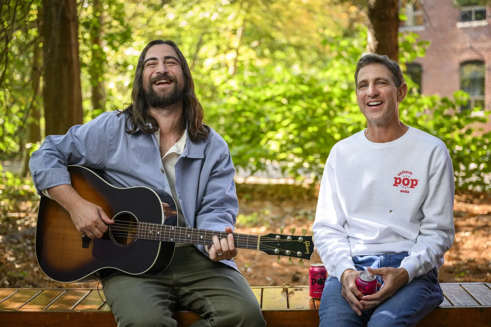 Two men sitting on a wooden bench outdoors, one playing a guitar and the other holding a soda can, both smiling and enjoying a sunny day in a park with green trees in the background.