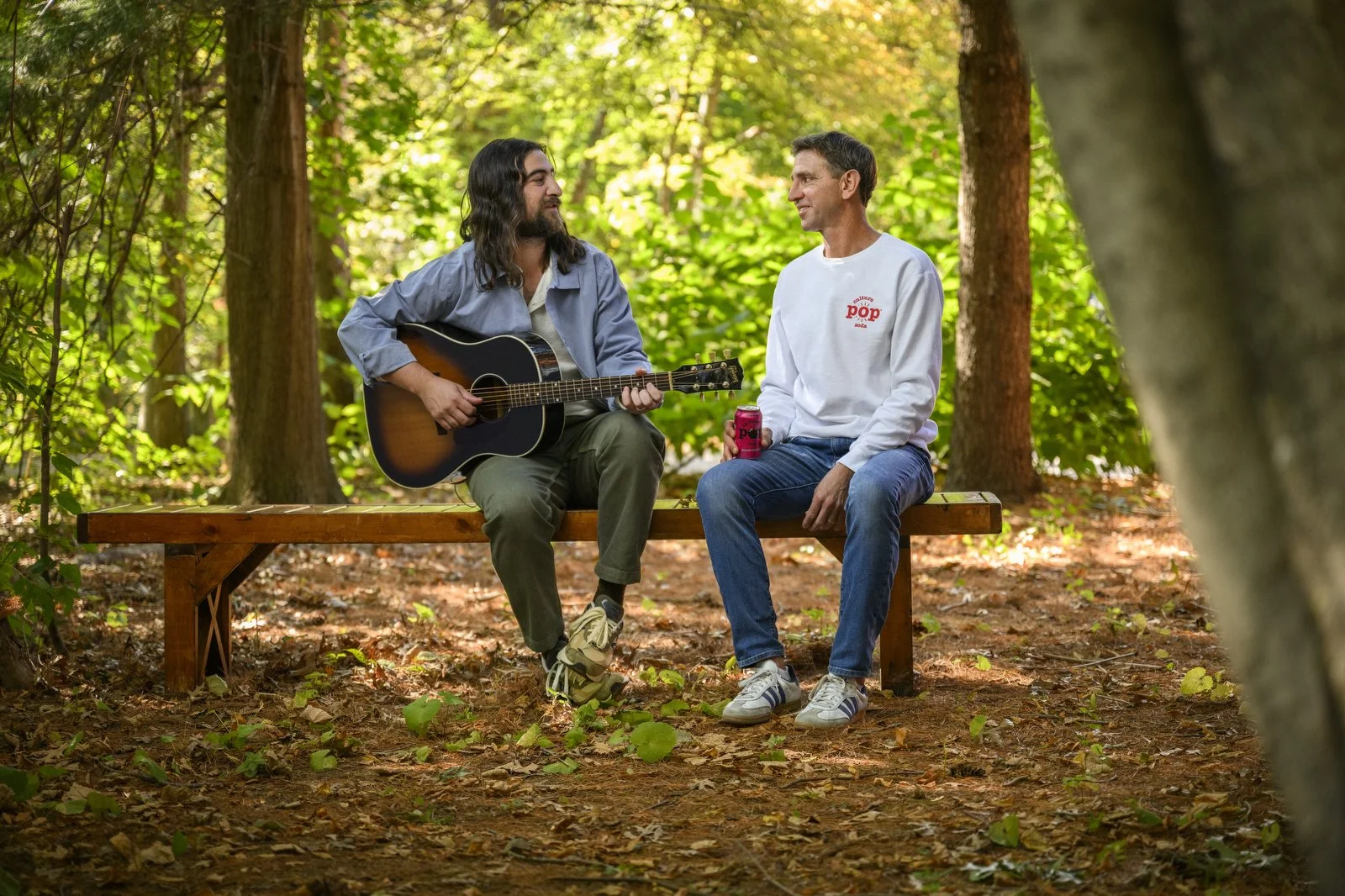 Two men sitting on a wooden bench in a forest. One is playing guitar while the other watches and listens. The man playing guitar has long dark hair and a beard, wearing a gray shirt and green pants. The other man, wearing a white sweatshirt and jeans, holds a can of soda. Sunlight filters through green leaves, creating a serene outdoor scene.
