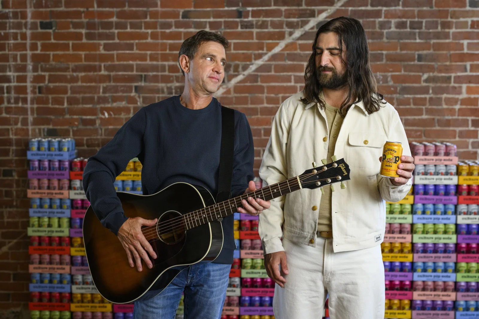 Two men standing in front of a colorful wall of cans, one holding a guitar and the other holding a can of pop soda, in front of a brick wall.