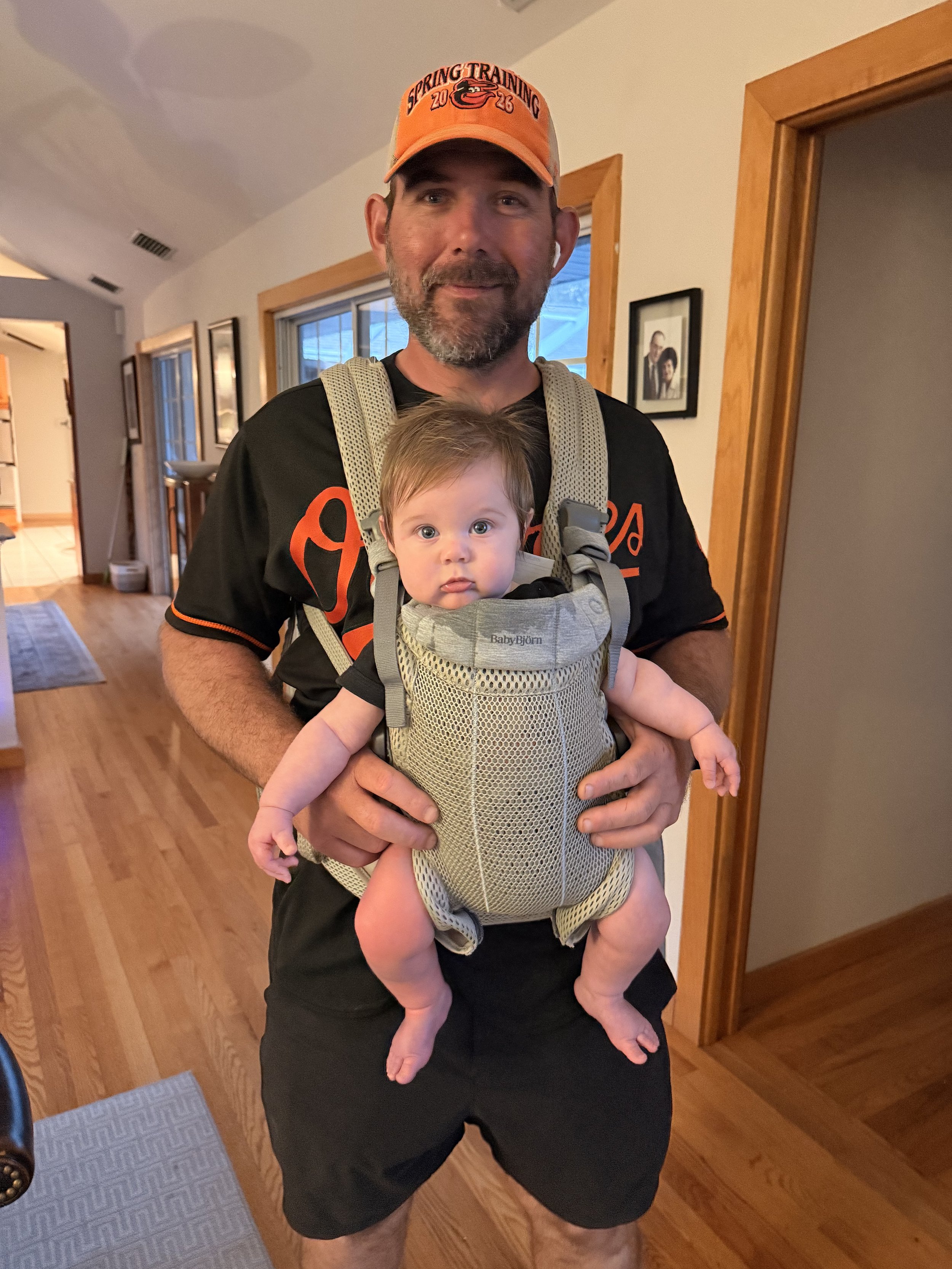 A man with a beard and short brown hair wearing a black t-shirt with orange graphics, black shorts, and an orange cap with 'Spring Training 2026' and a cartoon snake logo. He is holding a baby with blonde hair, blue eyes, and chubby cheeks, who is wearing a gray BabyBjörn baby carrier. The background shows a home interior with wooden floors, wall art, and a kitchen area.