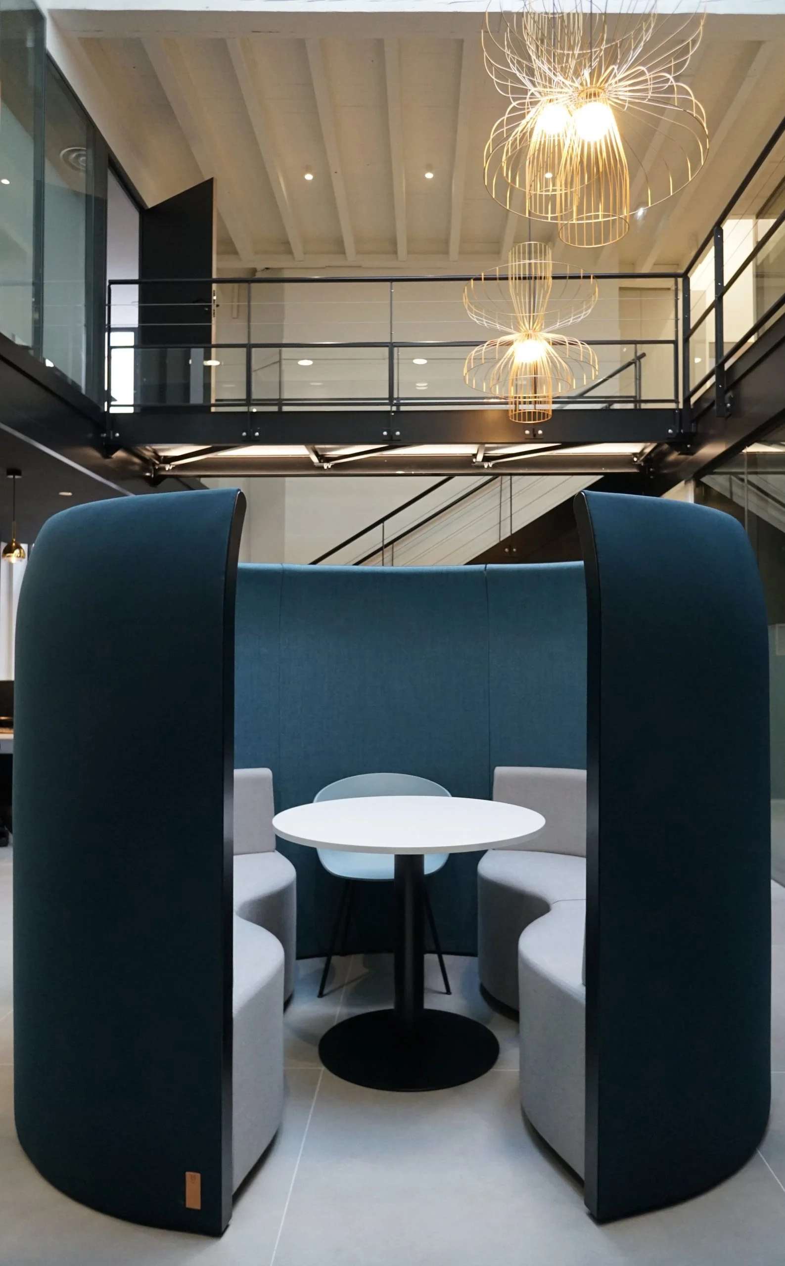 Une zone de détente ou de réunion dans un espace moderne, meublée de fauteuils blancs, entourée de panneaux acoustiques noirs, avec une petite table ronde et des chaises. Au-dessus, un escalier avec une rampe métallique et des luminaires suspendus au