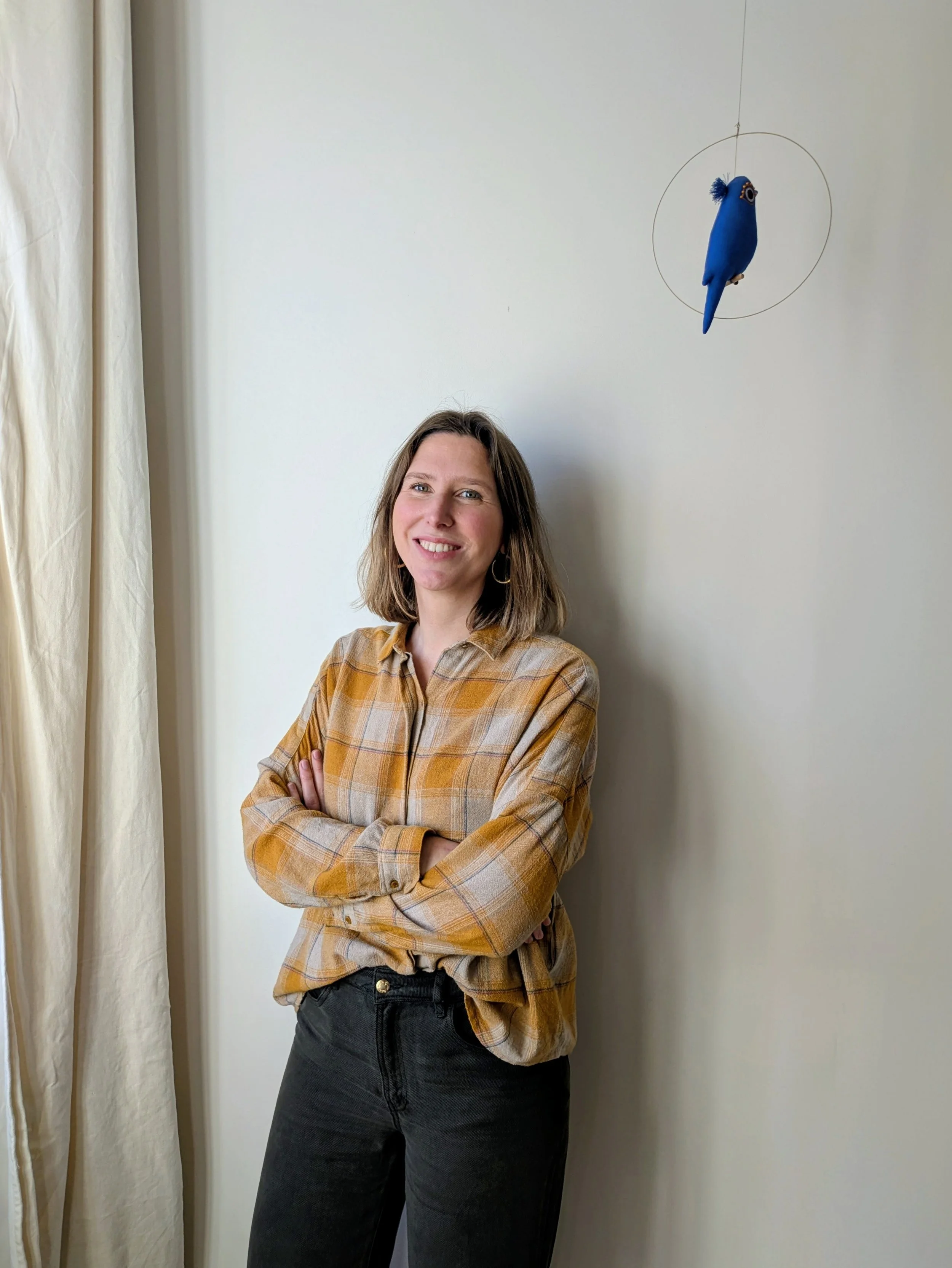 Une femme souriante portant une chemise à carreaux marron et noir, avec des bras croisés, se tient devant un mur clair avec un mobile en forme d'oiseau bleu suspendu au plafond, et une nappe beige à gauche.