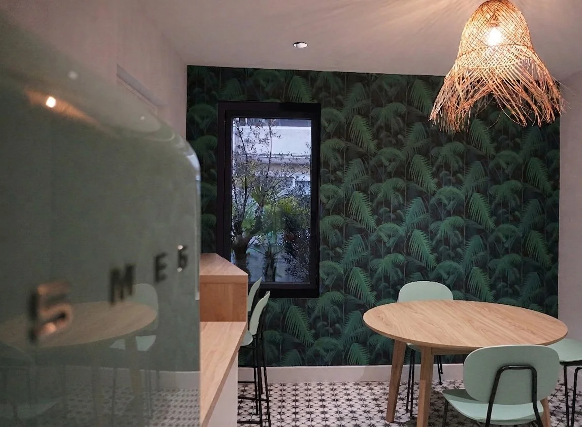 Une salle à manger avec un mur à motif feuillage végétal, une table ronde en bois entourée de chaises vert pastel, un éclairage suspendu en rotin, et une fenêtre transparente laissant entrer la lumière naturelle.