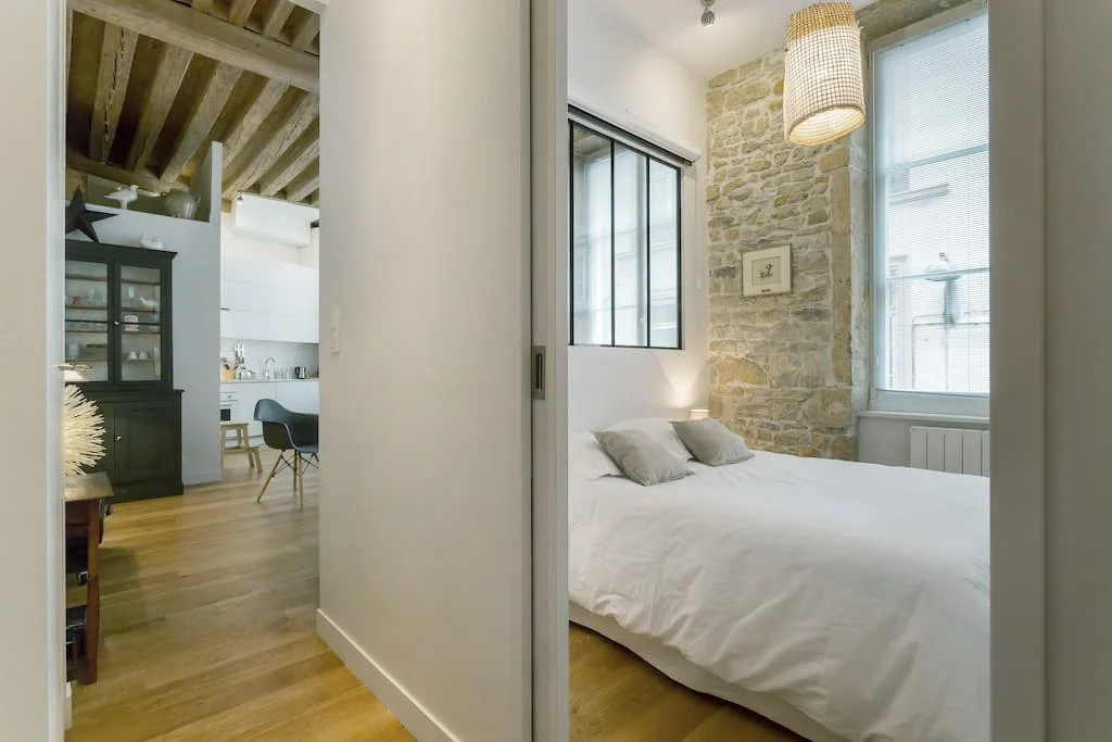 Une chambre avec un lit blanc, mur en pierre, grande fenêtre, lumières douces, style moderne et confortable.