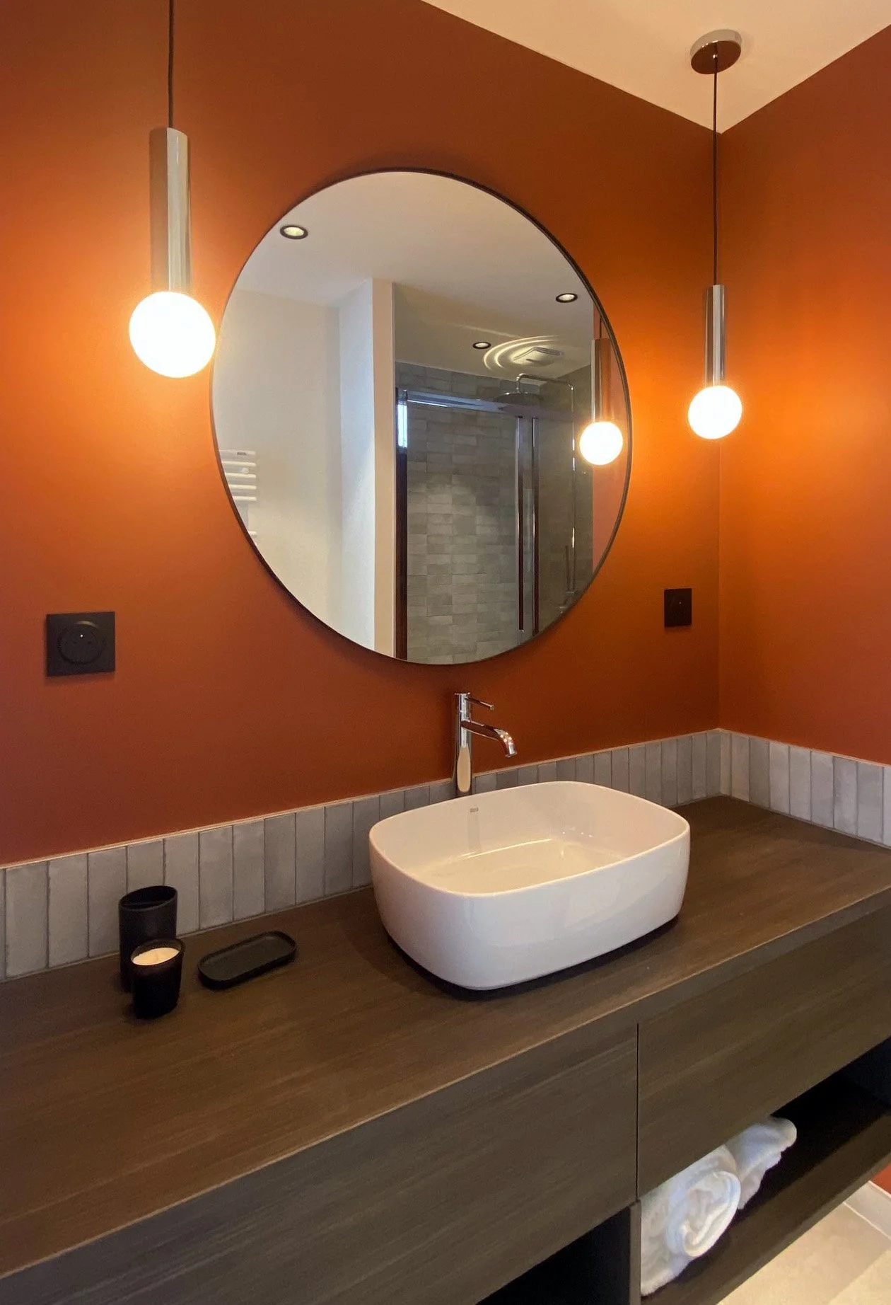 Un miroir rond au-dessus d'un lavabo blanc dans une salle de bain avec murs orange. Suspensions lumineuses de chaque côté du miroir. Vanités en bois avec des objets noirs et des bougies, un lavabo et des serviettes blanches en dessous.