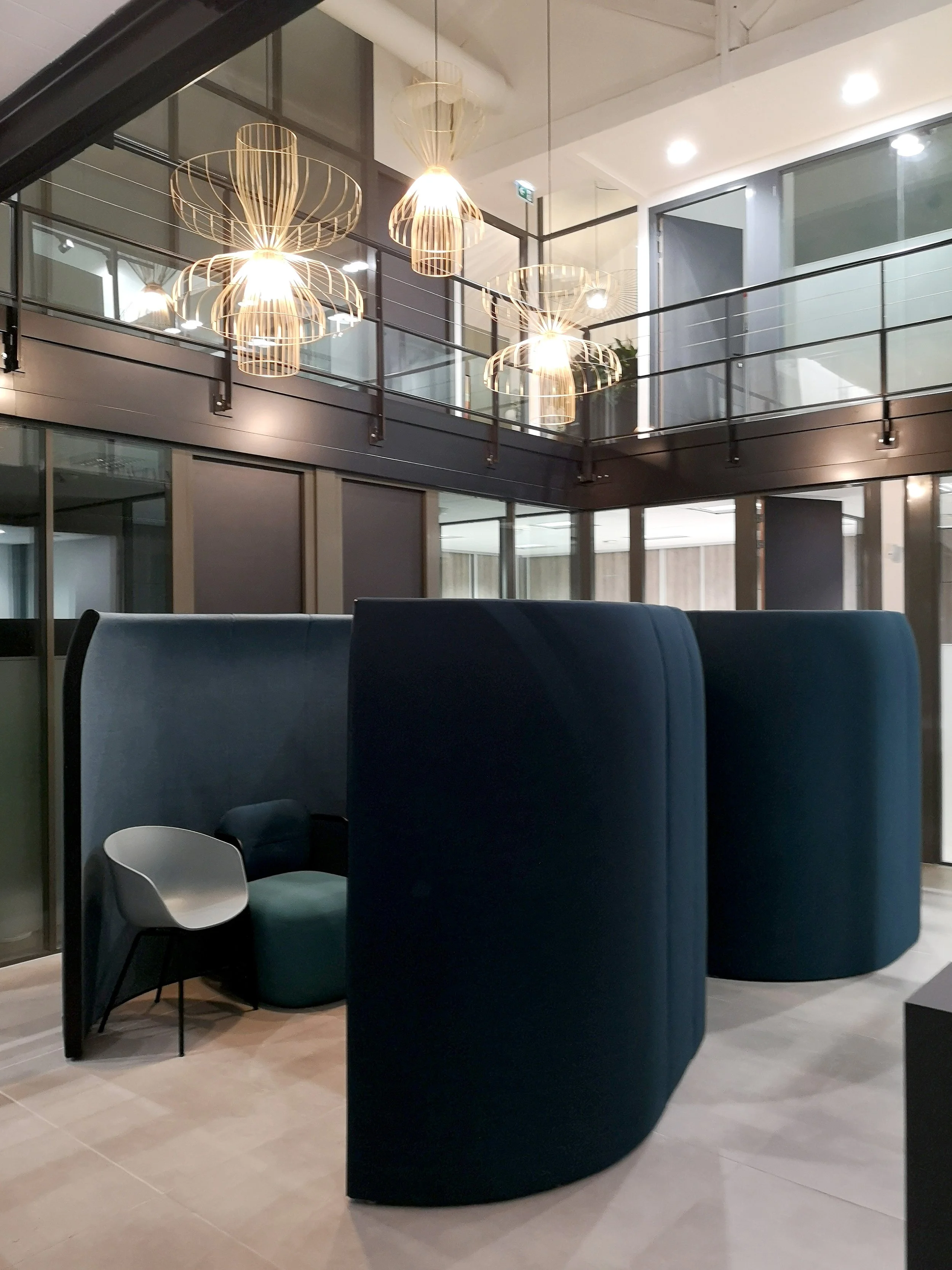 Intérieur d'un espace moderne avec un mur en verre, un plafond blanc, des lampes suspendues en forme de cage en métal, des chaises et un fauteuil dans un coin.
