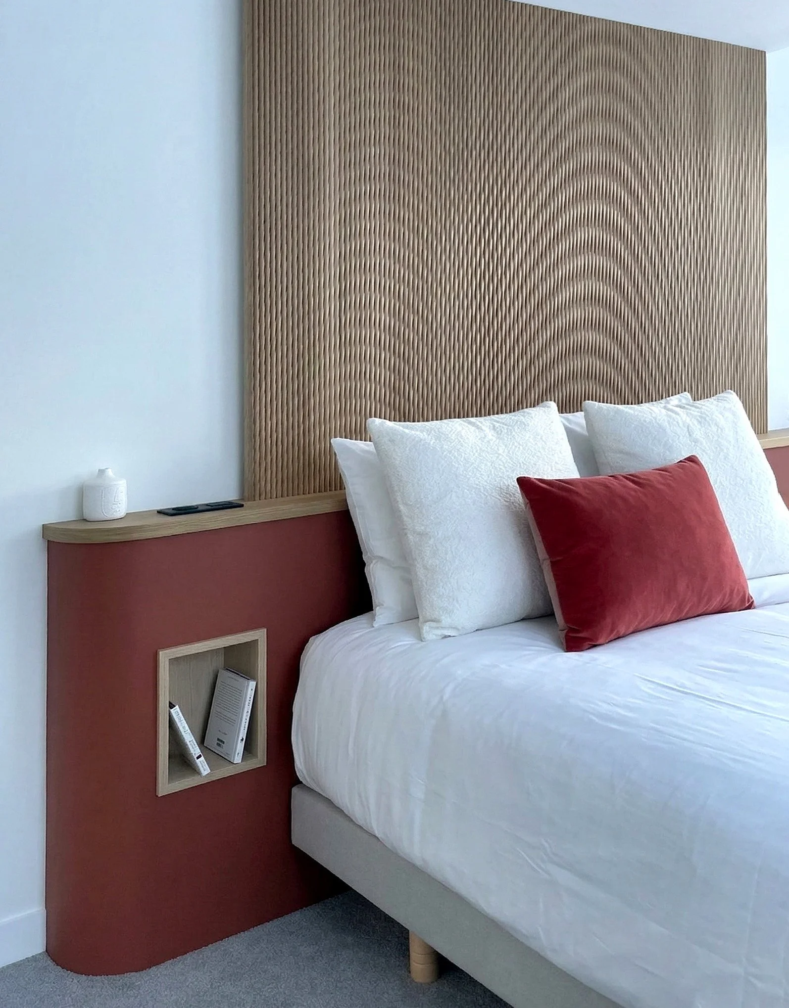 Chambre d'hôtel avec un grand lit, un tête de lit en bois avec un motif en relief, deux coussins blancs et un coussin rouge sur le lit, une petite table de chevet avec une lampe et des livres, et un mur blanc.