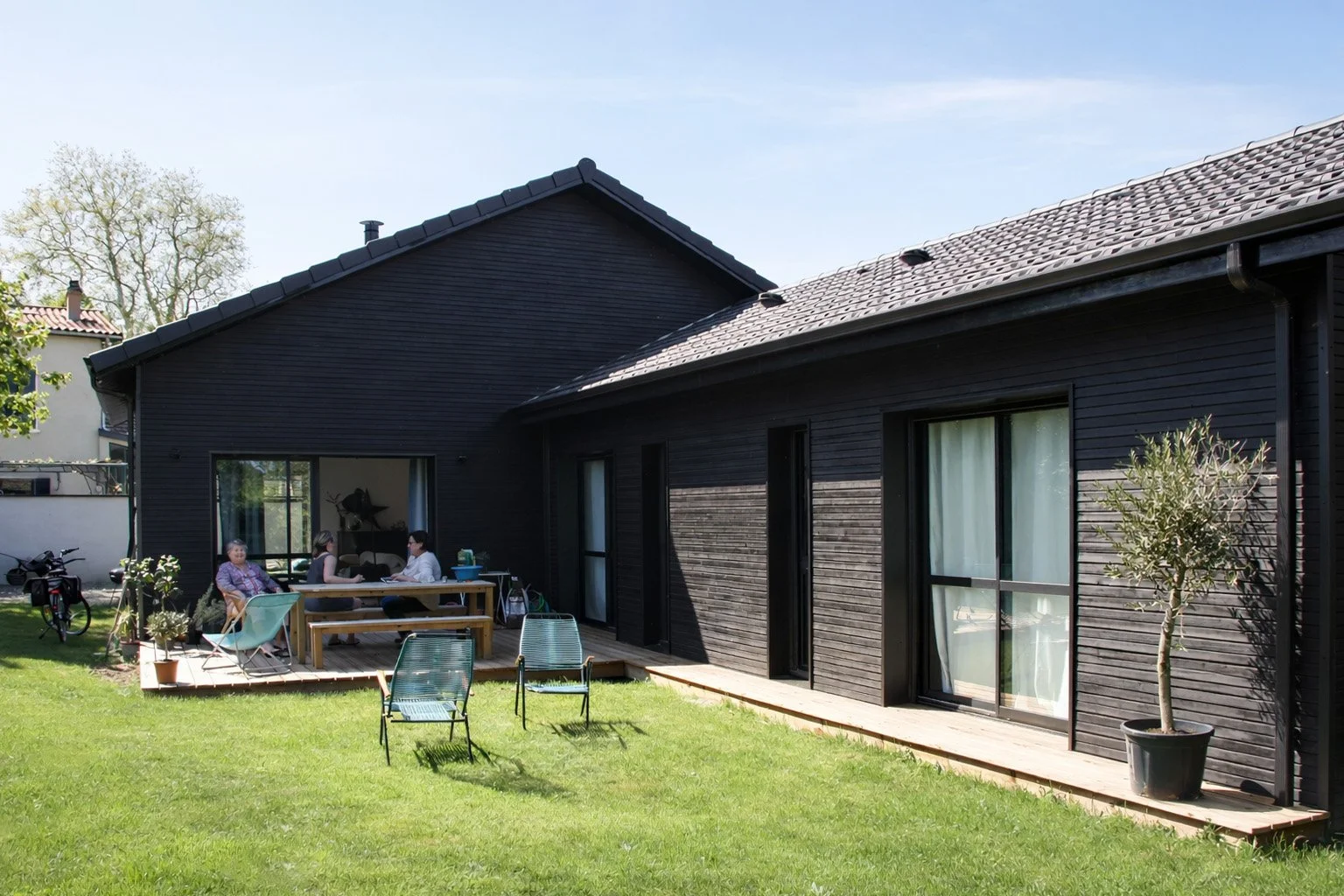 Une maison moderne en bois noir avec une terrasse en bois, quelques personnes assises à une table, un vélo et un petit arbre en pot dans le jardin.